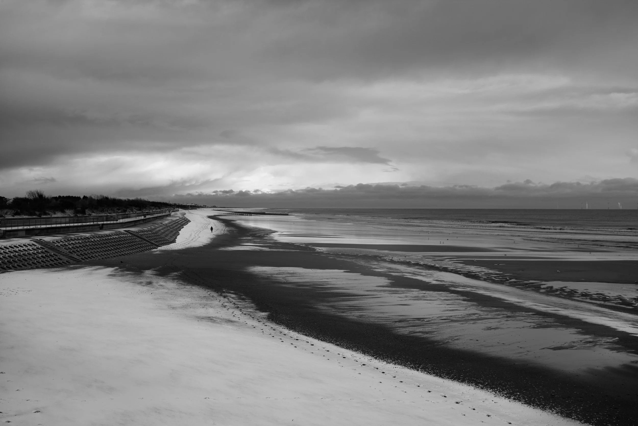 Black and white photo of a beach with sand and water, cloudy sky, and a person walking in the distance.