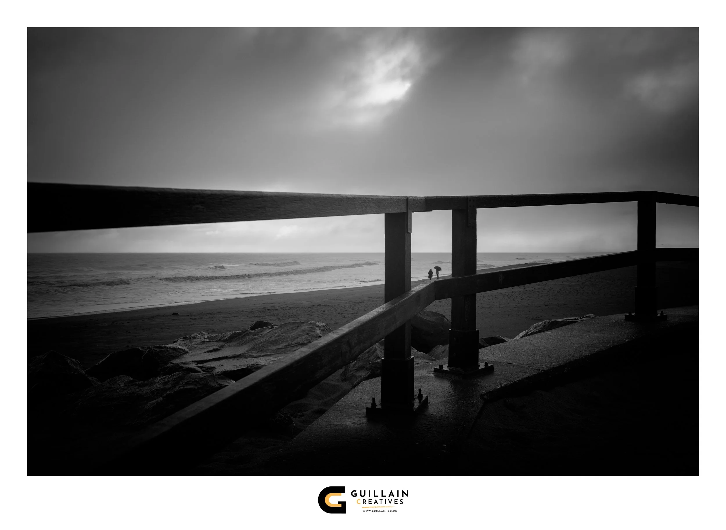 Black and white photo of a beach with a wooden railing, two people walking along the shoreline, clouds in the sky, and ocean waves.