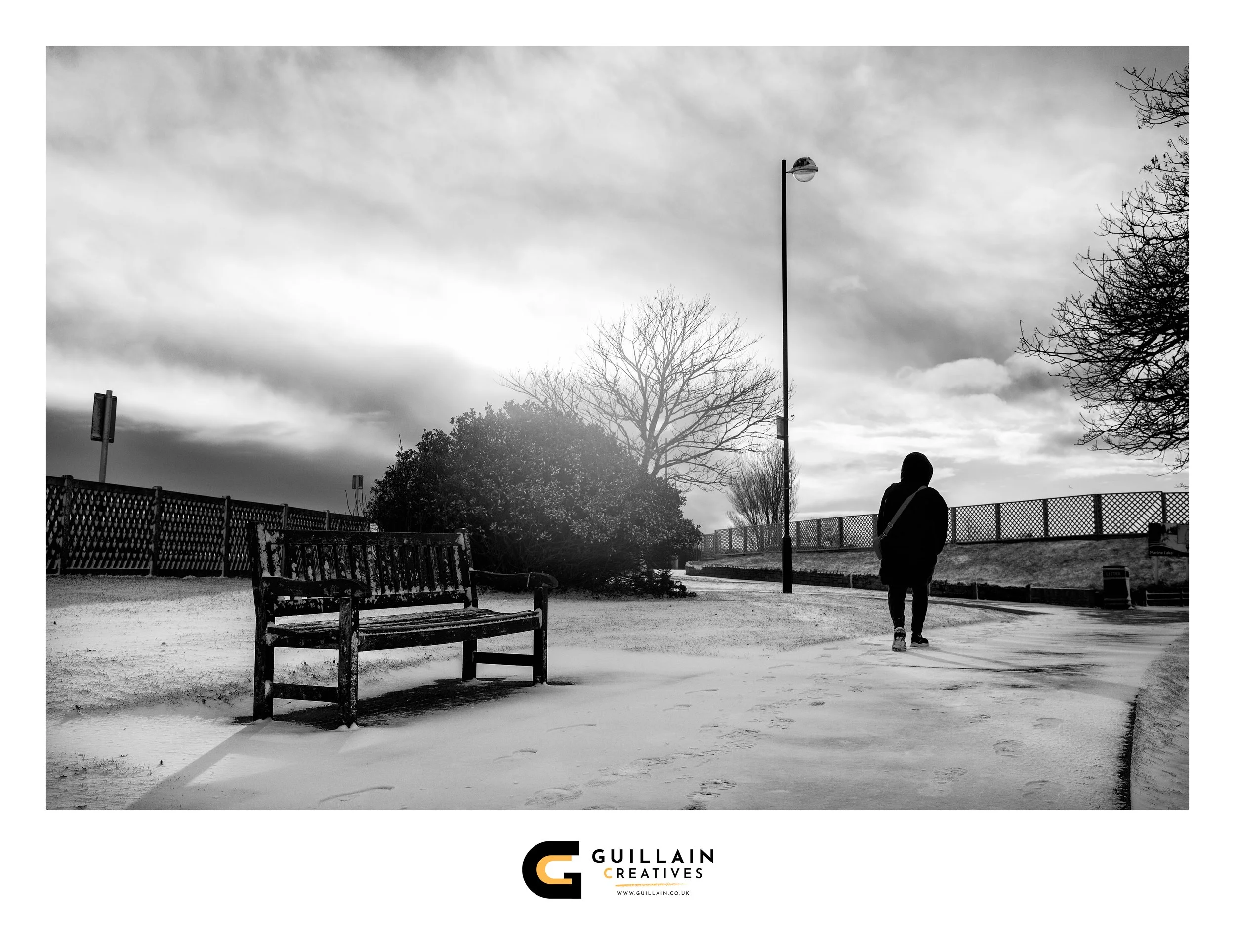 A person dressed in dark clothing walks on a snow-covered path in a park, with leafless trees, a wooden bench, and a lamppost visible. The sky is cloudy and overcast.
