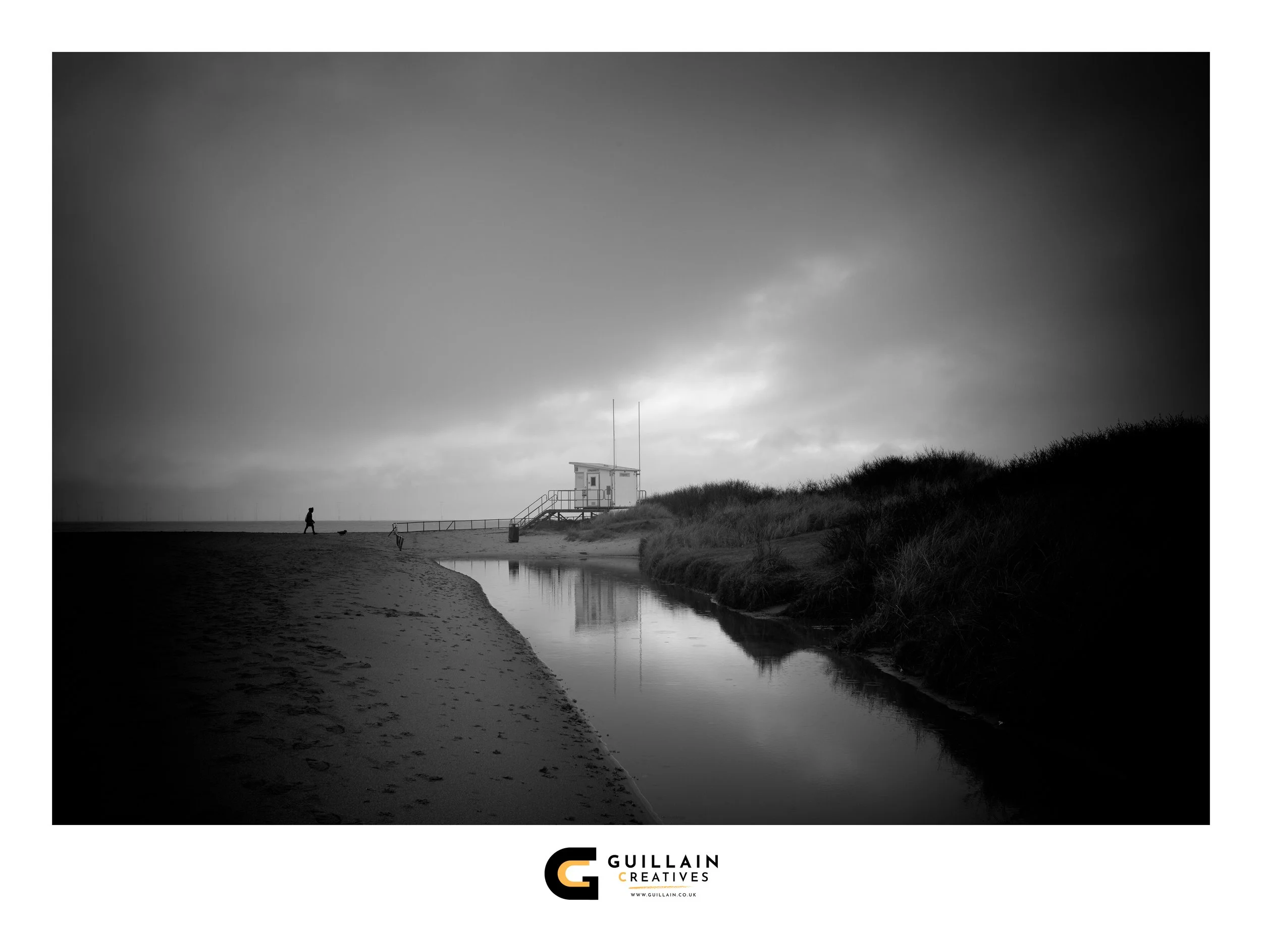 A black and white photo of a beach with a person walking a dog near the water, a lifeguard station on stilts, and grassy dunes, under a cloudy sky.