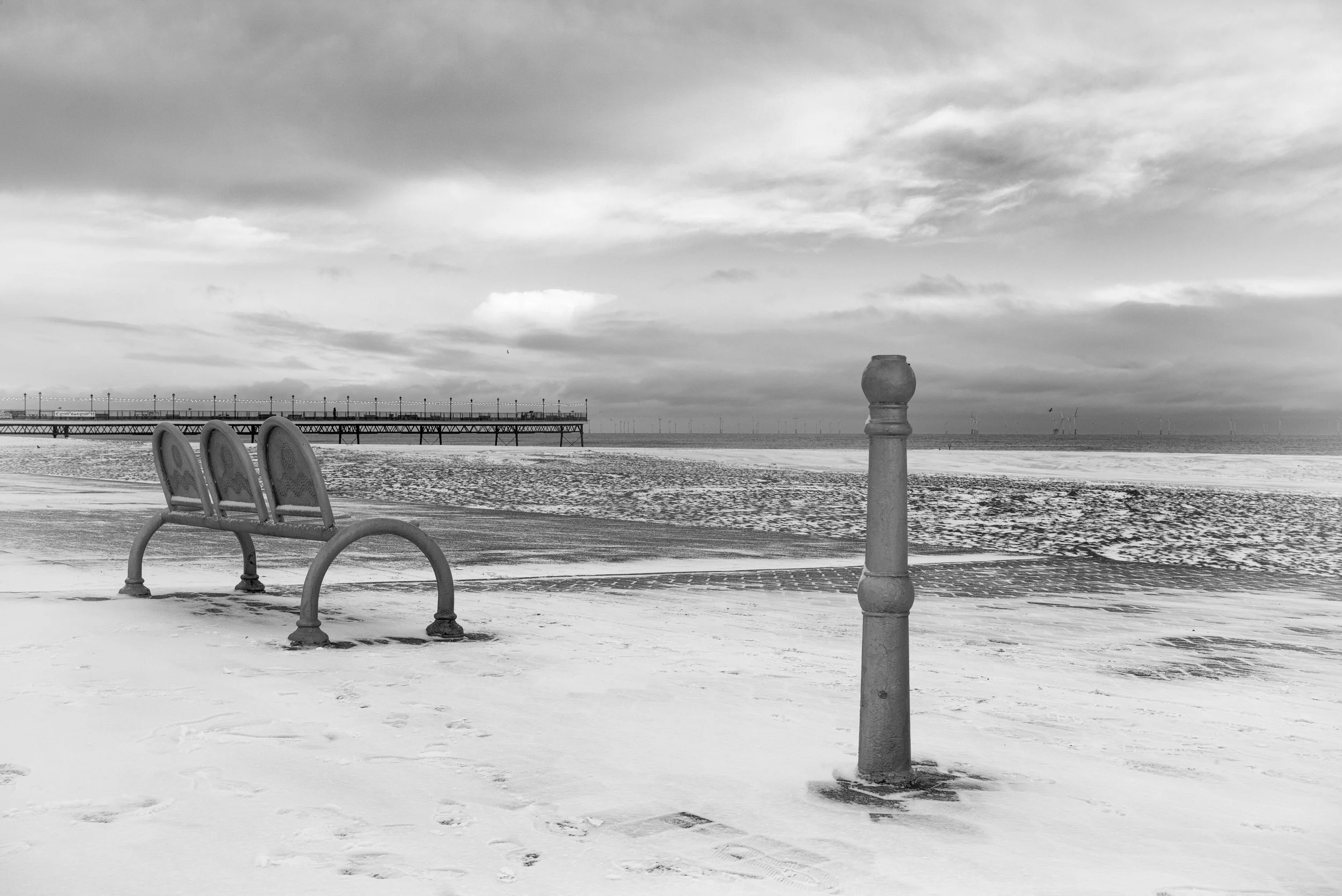 A black and white photo of a beach with three empty benches facing the ocean, a lamppost, a pier in the background, and wind turbines on the horizon, under a cloudy sky.