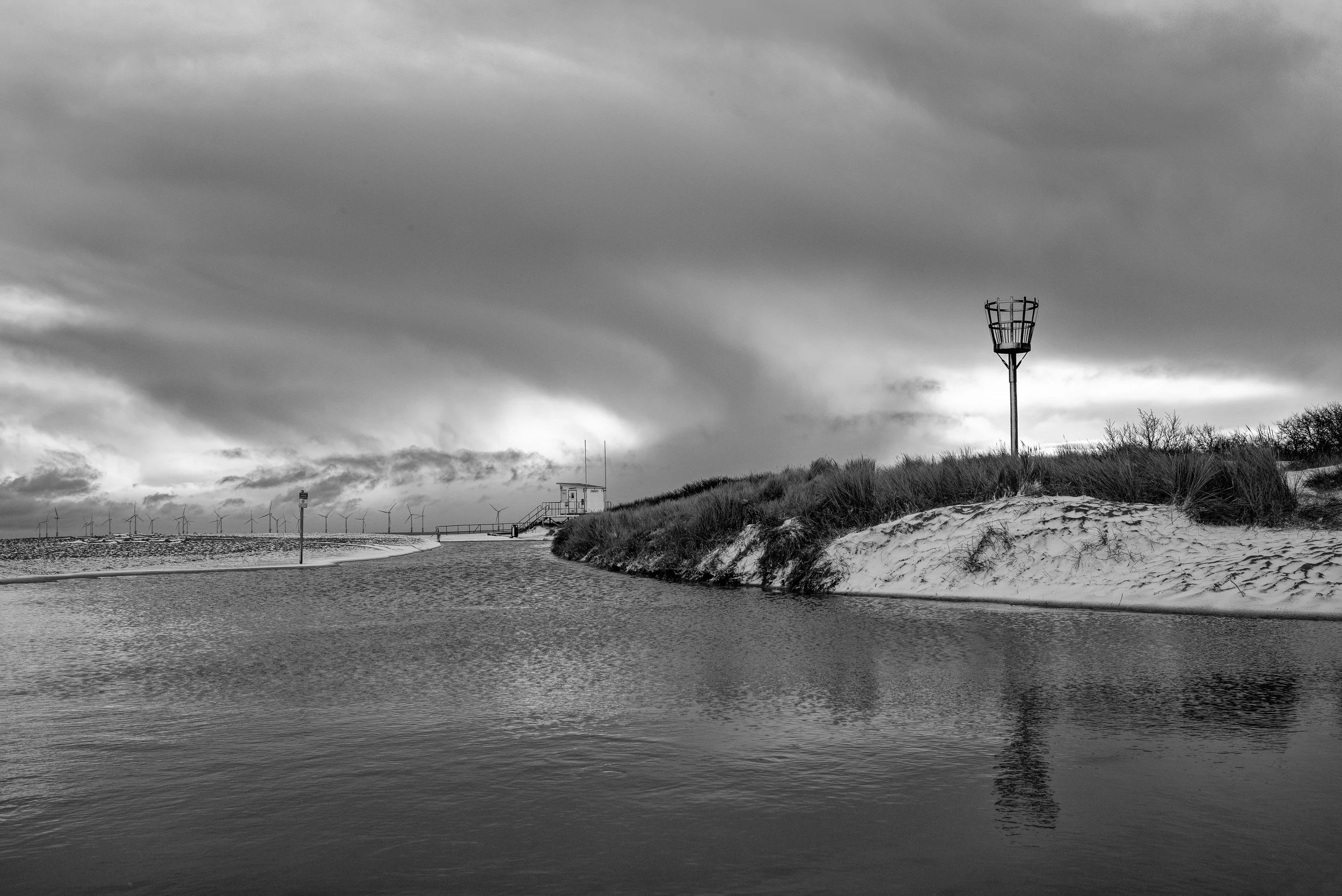 Black and white photo of a river or inlet with sandy and grassy banks, a navigation light, and wind turbines in the distance under a cloudy sky.