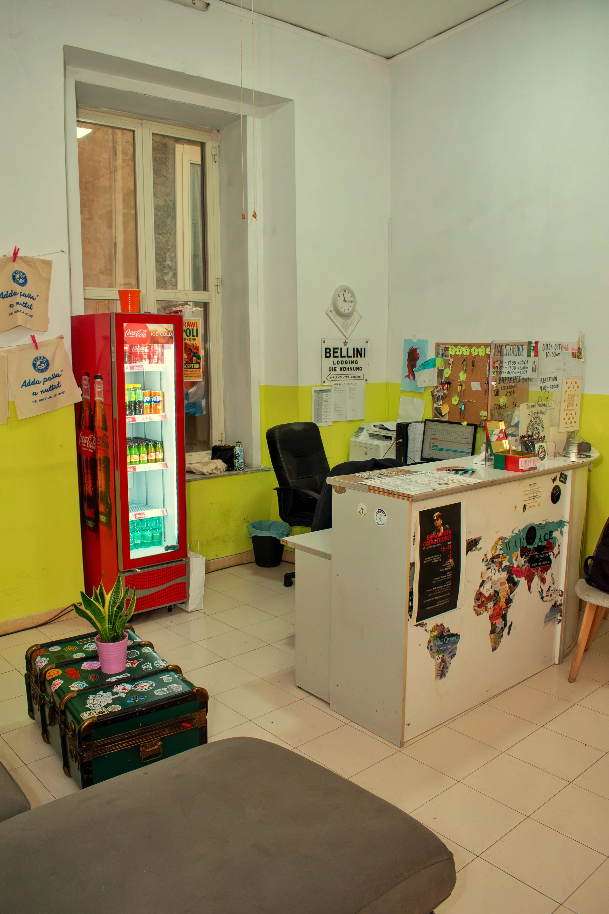 An office with a white desk featuring world map stickers, black office chair, black swivel chair, window with a ledge, red Coca-Cola mini fridge, and various decorations including a potted plant and posters.