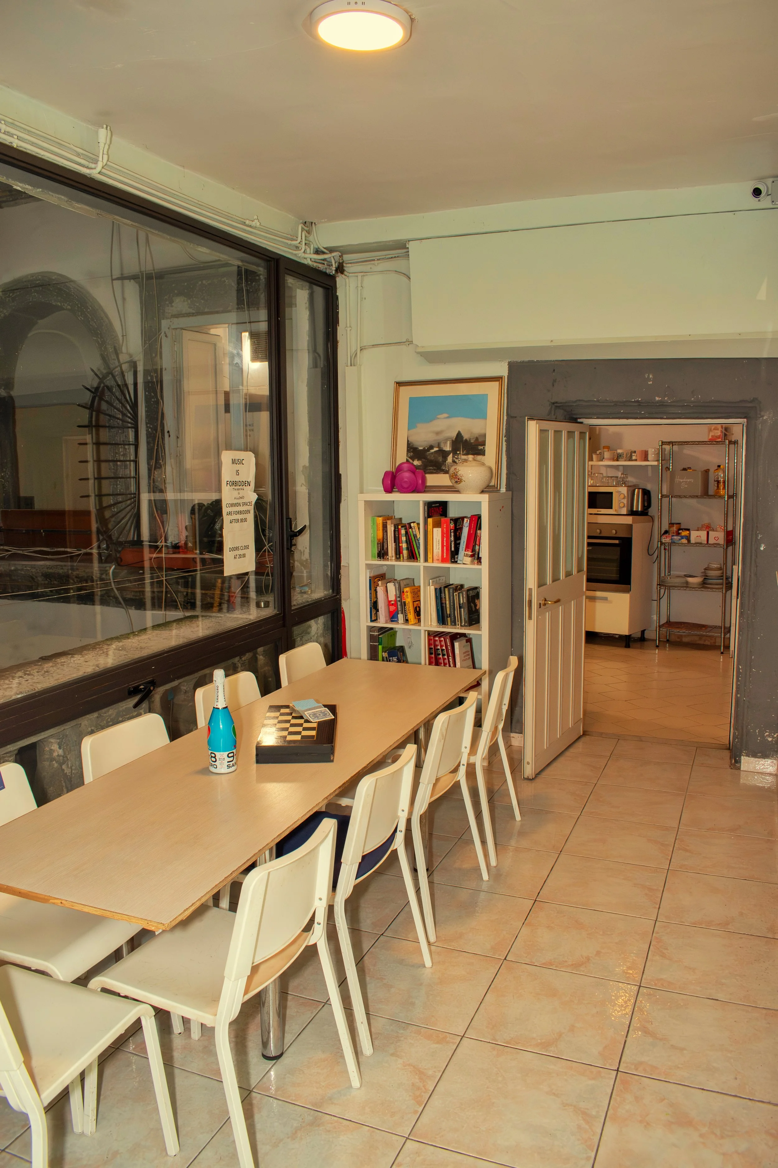 Interior of a dining area with a rectangular table, white chairs, a bookshelf with books, and a kitchen visible through an open door, with gray and white walls and tiled floor.