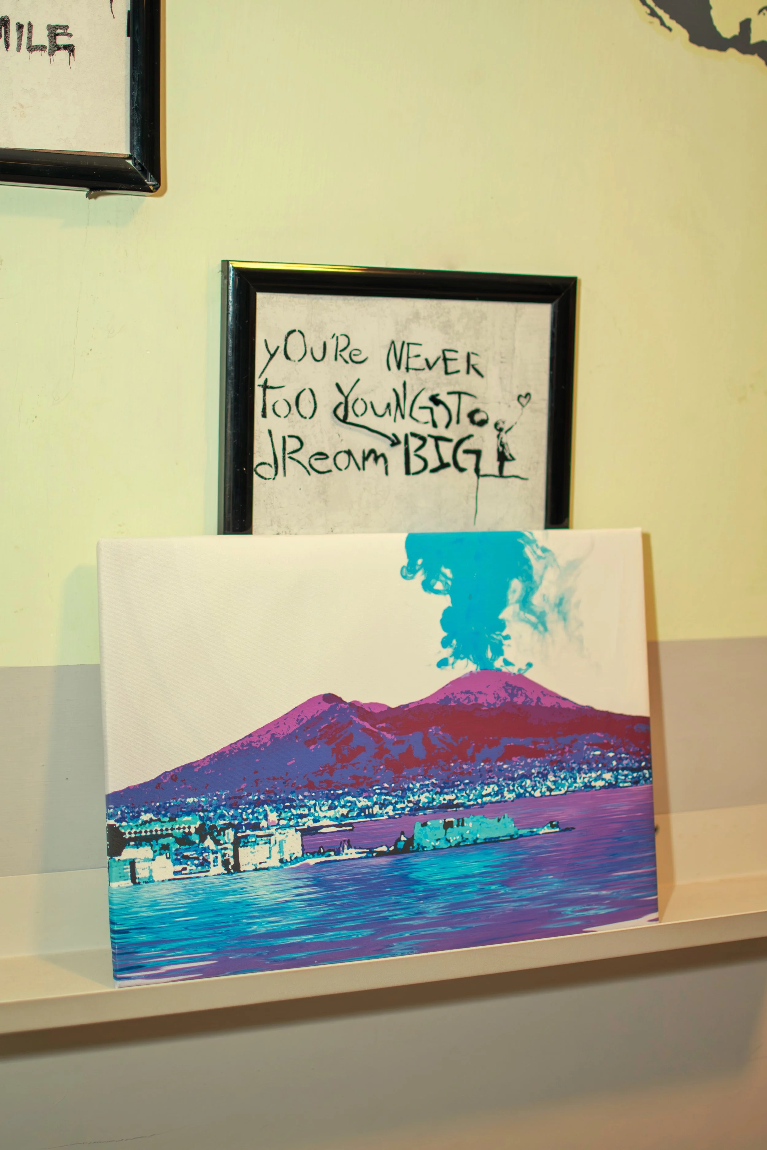 Colorful painting of a volcano emitting blue smoke, with water and buildings in the foreground, placed on a shelf in front of two framed quotes on the wall.