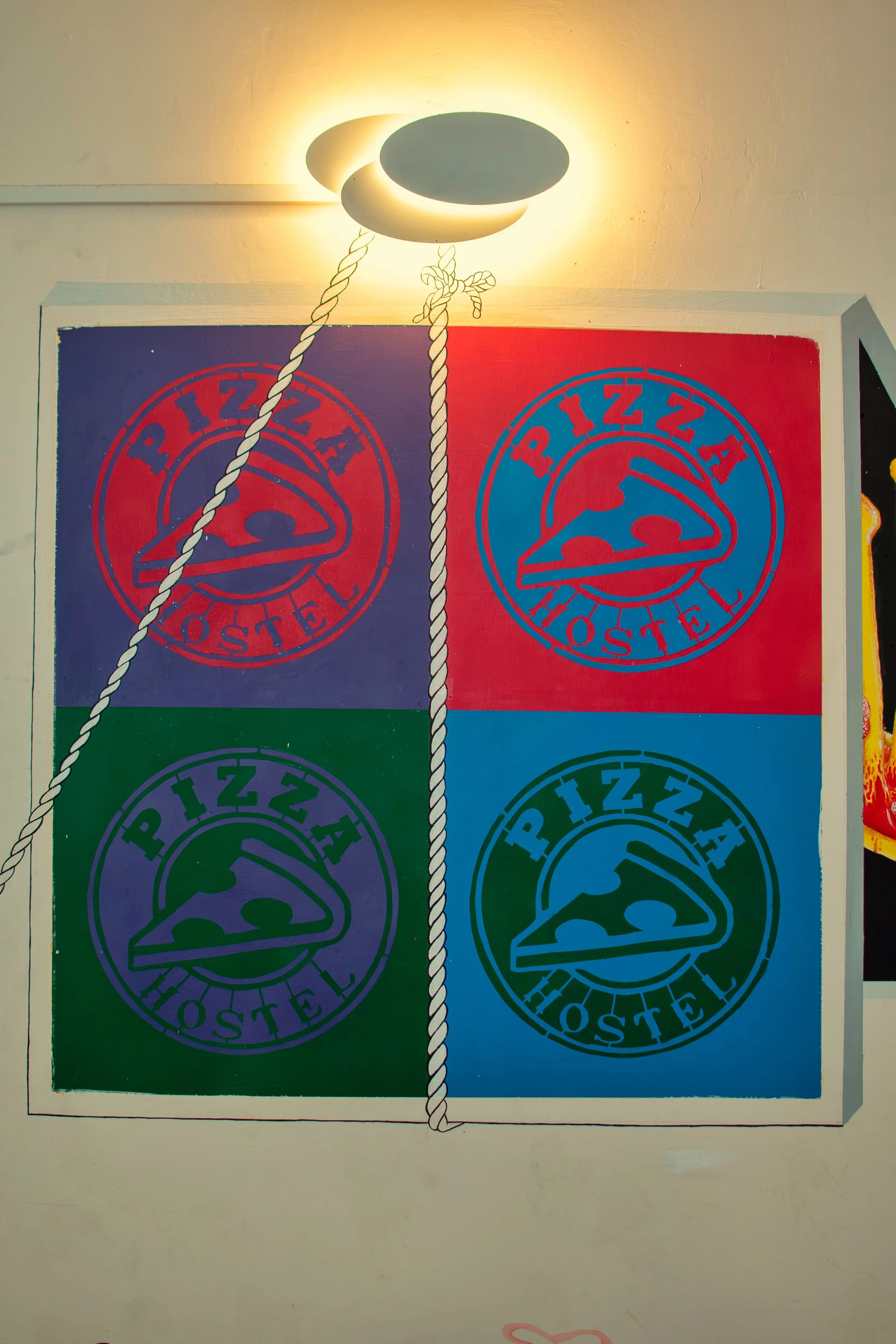 Colorful wall art with four quadrants, each featuring a pizza logo with the words 'Pizza Hostel'; the quadrants are colored purple, red, green, and blue.