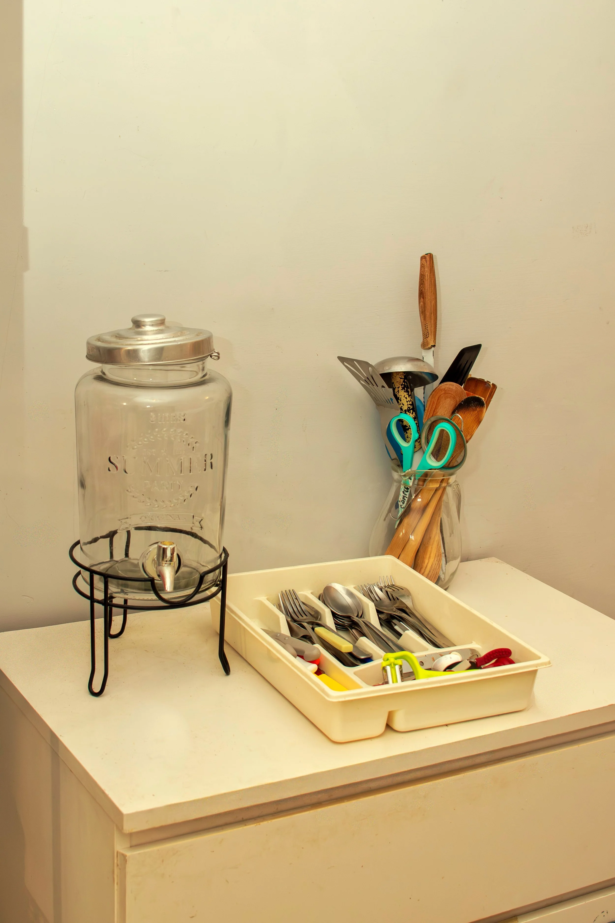 A counter with a glass beverage dispenser, a white utensil tray with silverware, and a jar holding various cooking utensils such as knives, scissors, and wooden spoons.