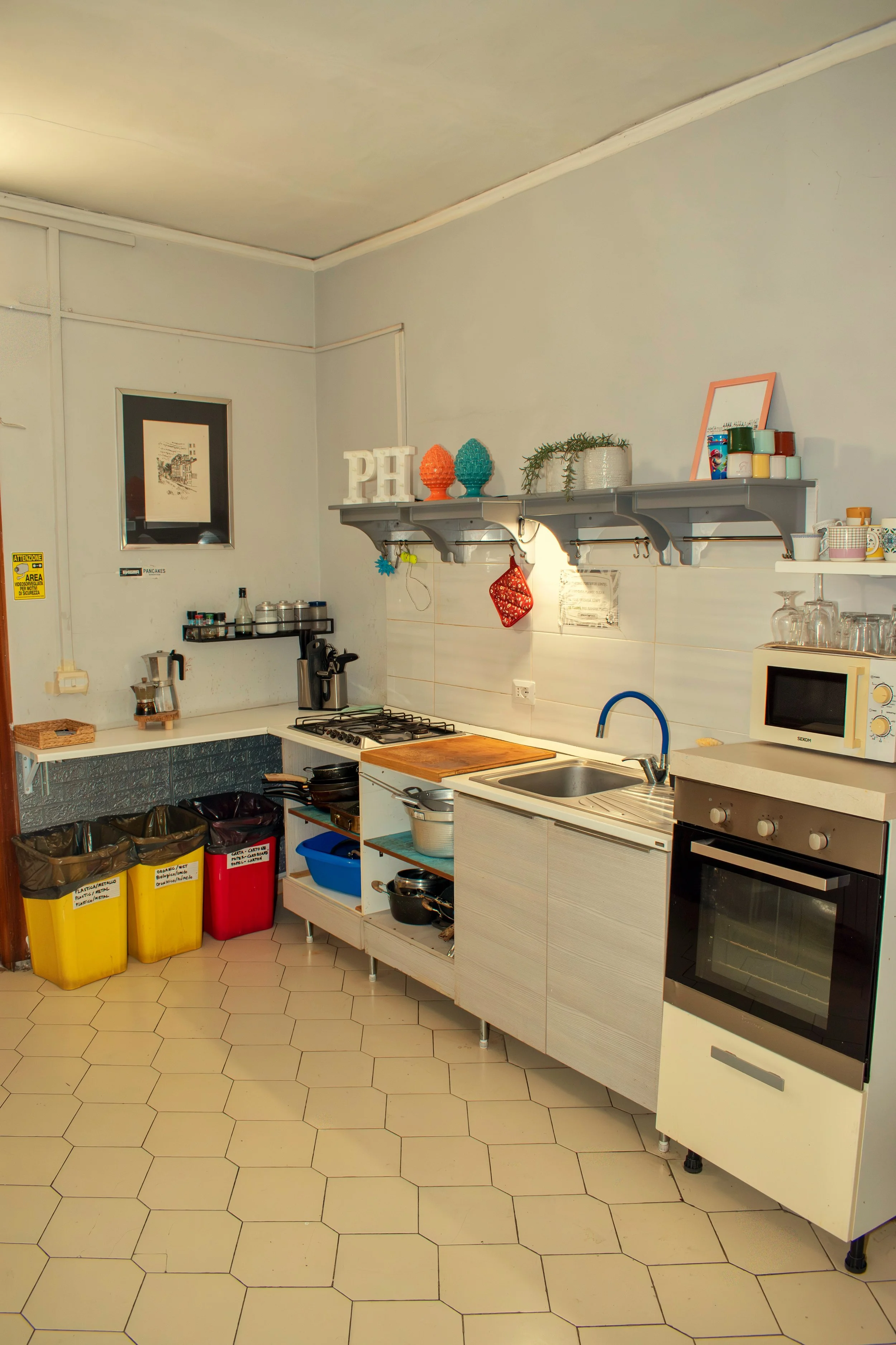 A small kitchen with a white counter, a stove, a microwave, and a black oven. Open shelving with decorative items, kitchen supplies, and glasses. Trash bins under the counter and a tiled hexagon floor.