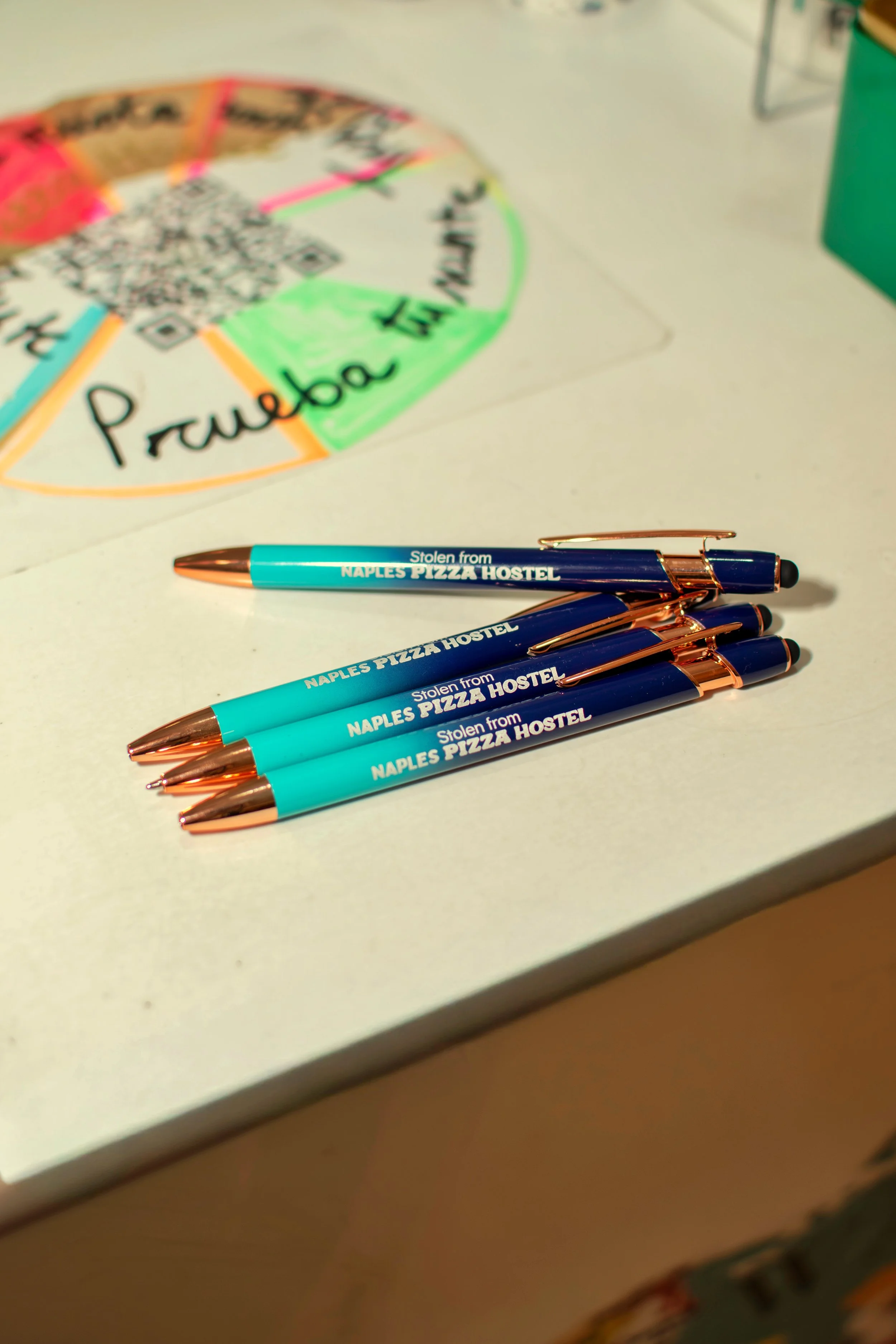 Four blue pens with rose gold accents, labeled "Stolen from NAPLES PIZZA HOSTEL," are arranged on a white table next to a colorful life wheel drawing.