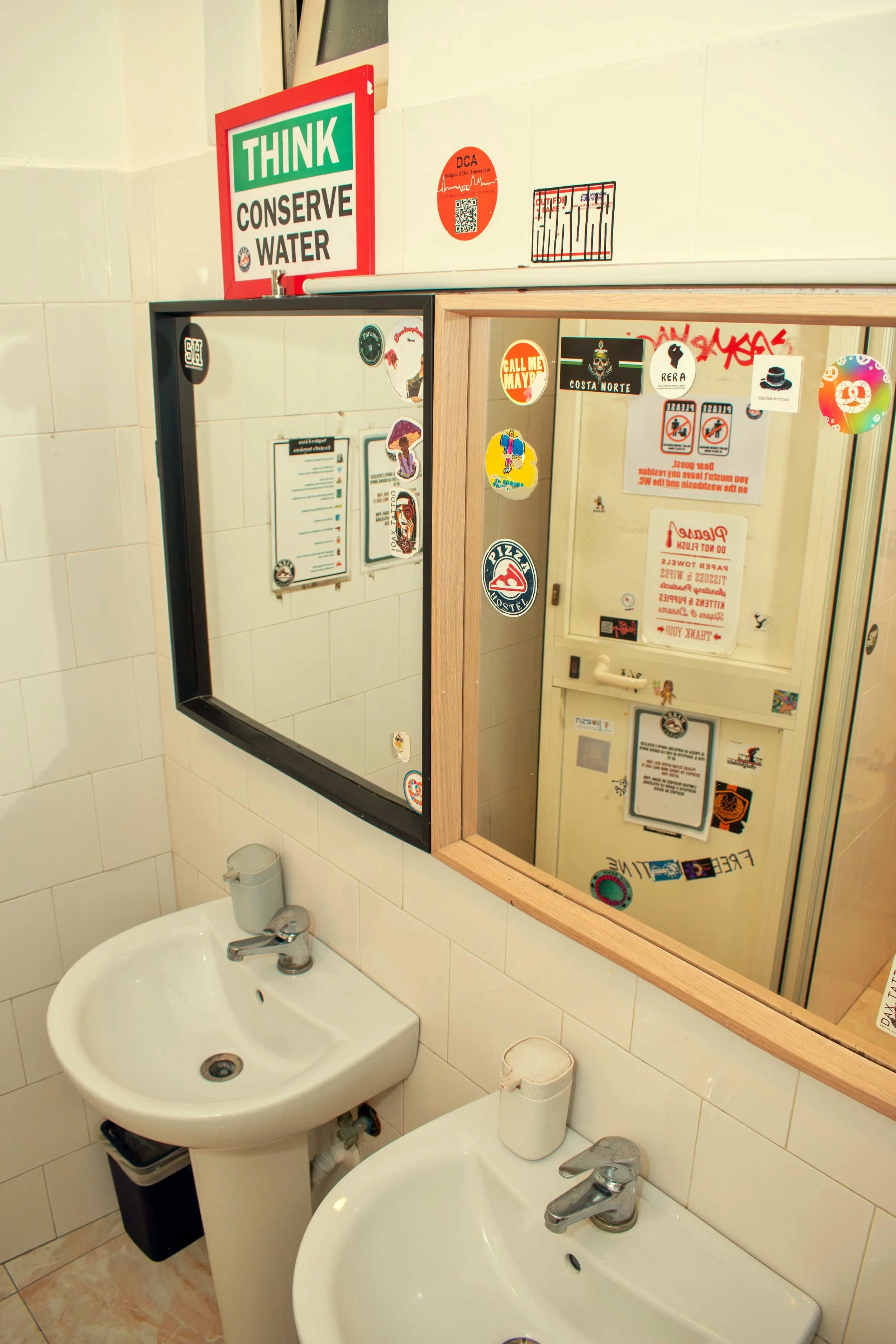 A bathroom with two white sinks, a mirror with colorful stickers, and a wall with various signs and stickers.