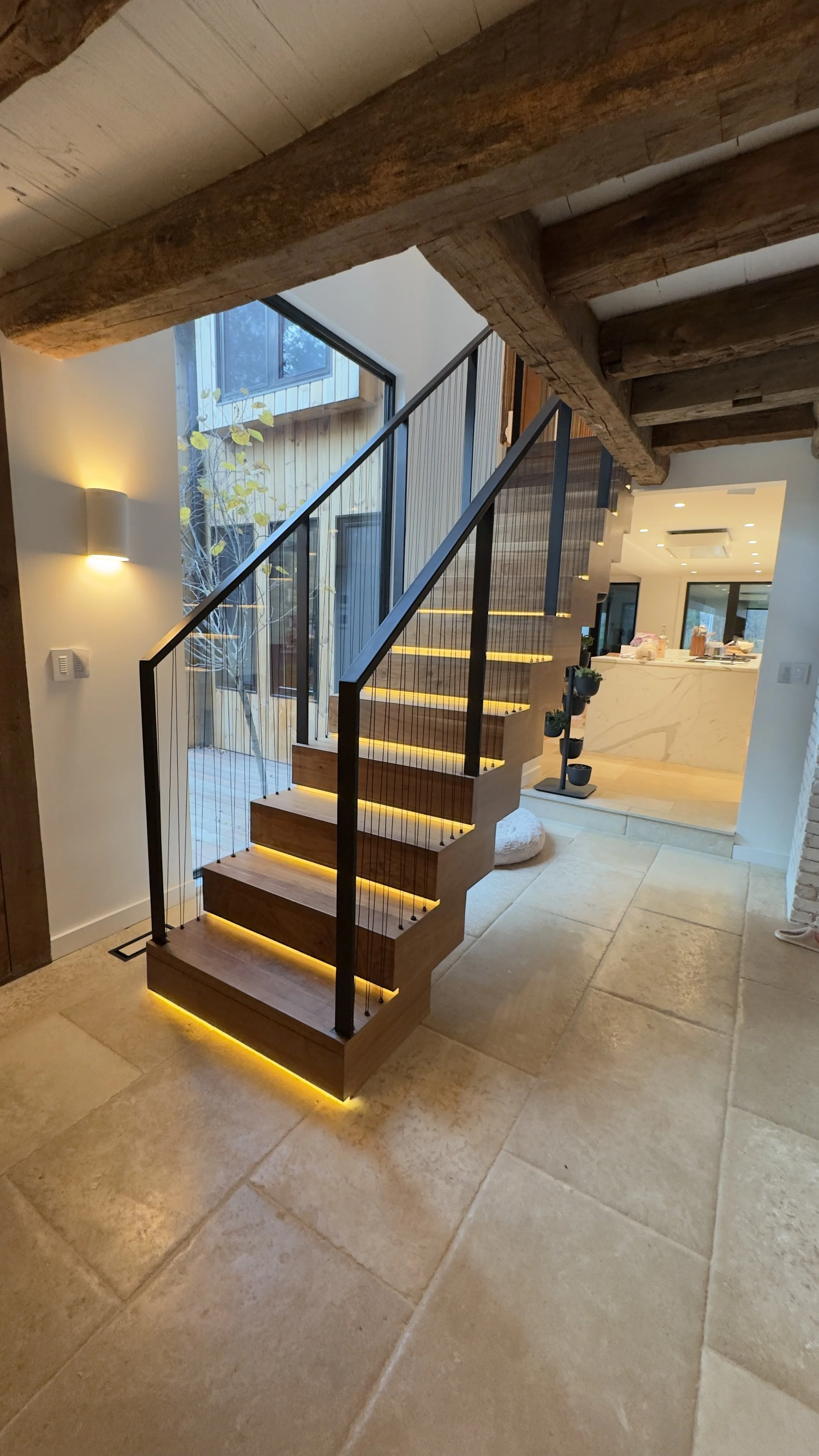 Modern interior staircase with illuminated risers, wooden treads, a black metal railing, and large windows letting in natural light in a contemporary home.