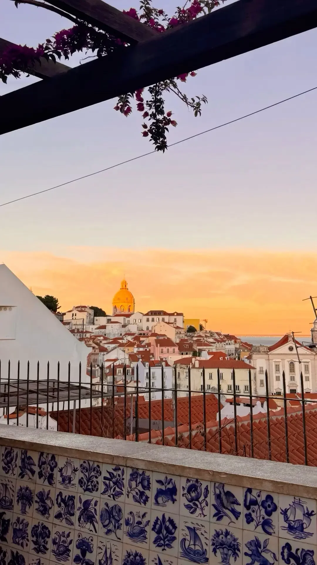 Vista de uma cidade com prédios de telhados vermelhos durante o pôr do sol, com um céu alaranjado ao fundo e uma cúpula dourada ao longe, visto de uma varanda com telhas de azulejo azul e cerca de ferro.