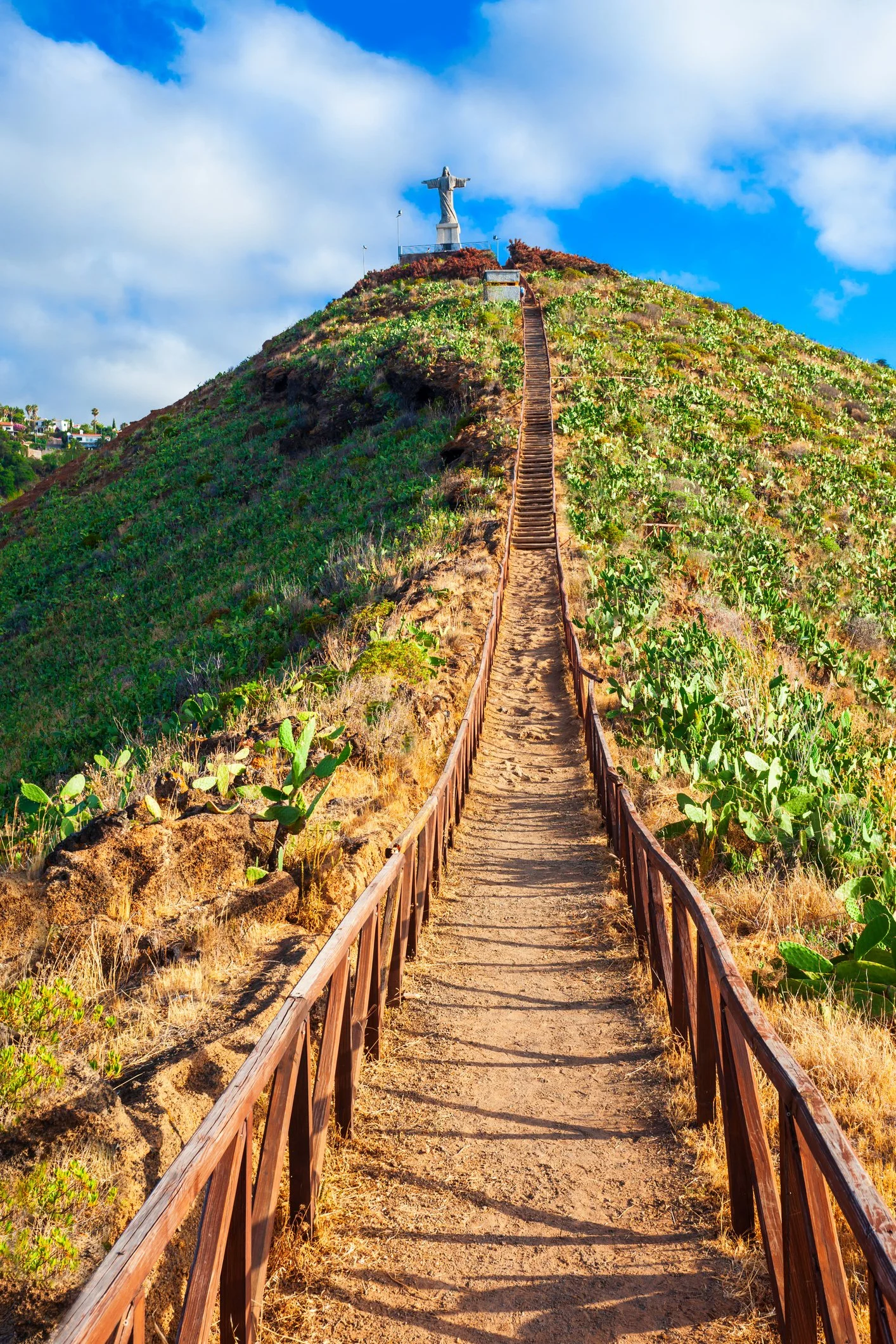 Caminho de terra com corrimãos de madeira que sobe uma colina até uma estátua de Cristo. Vegetação seca e cactus ao redor, céu azul com algumas nuvens.
