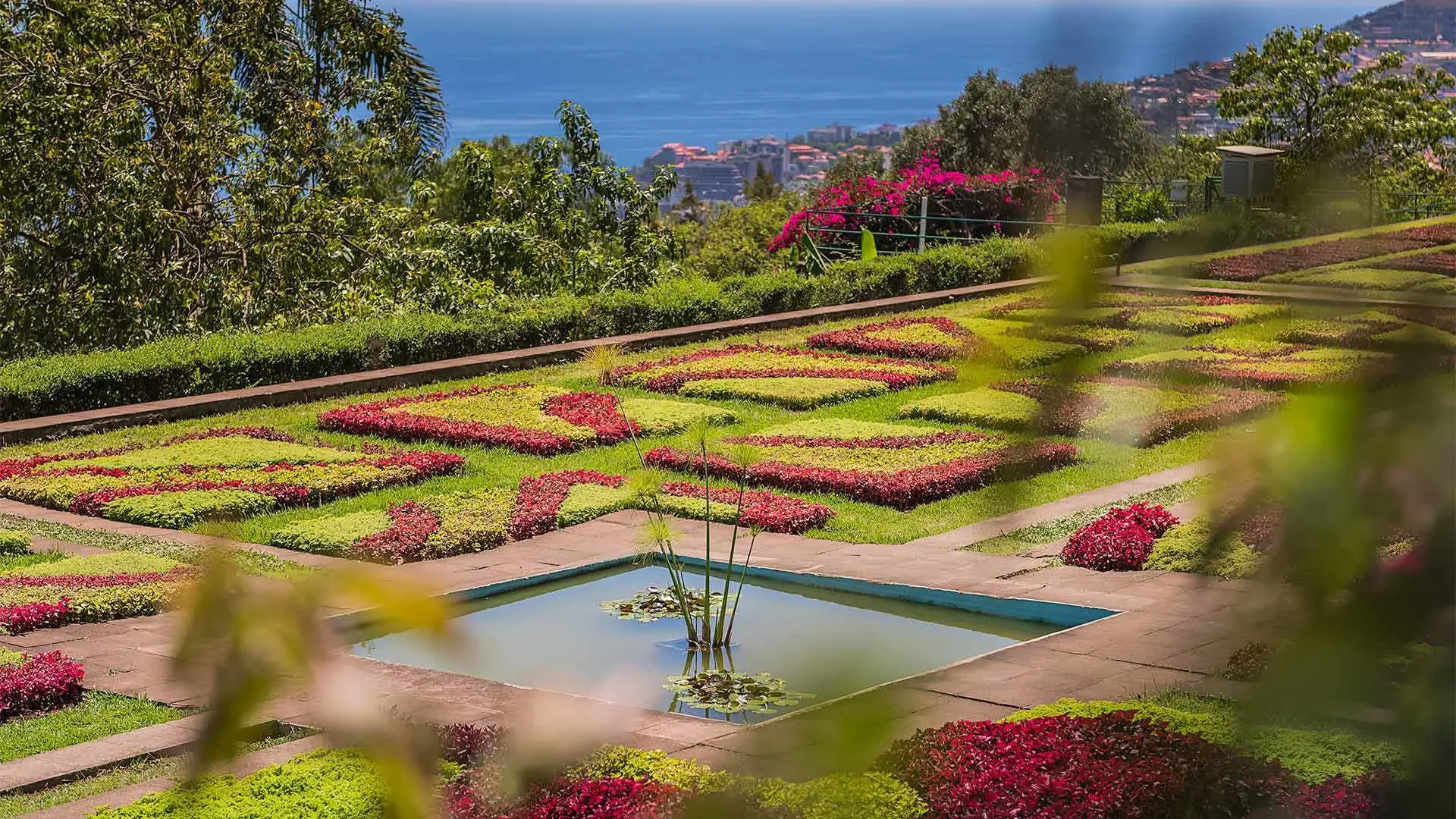 Jardim com canteiros de flores coloridas e uma pequena fonte de água ao centro, com vista para o mar ao fundo.