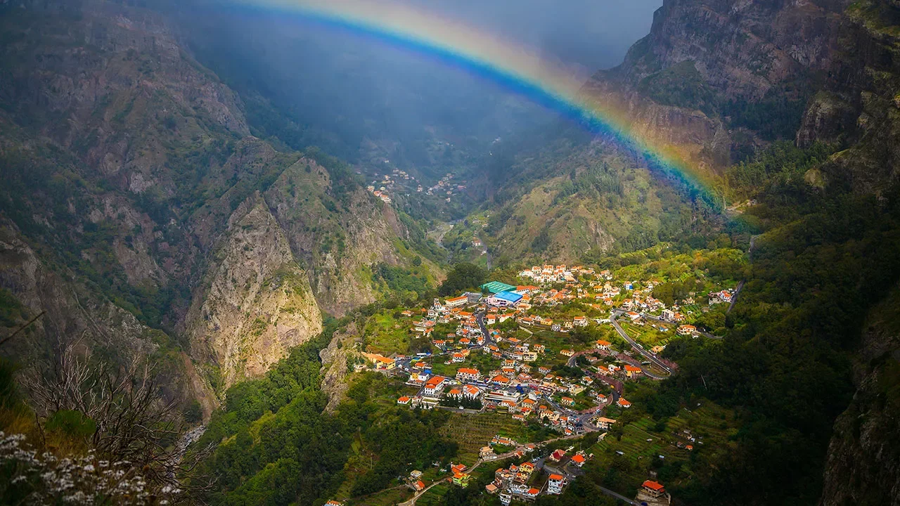 Paisagem de uma vila em um vale cercado por montanhas com um arco-íris no céu ao fundo.