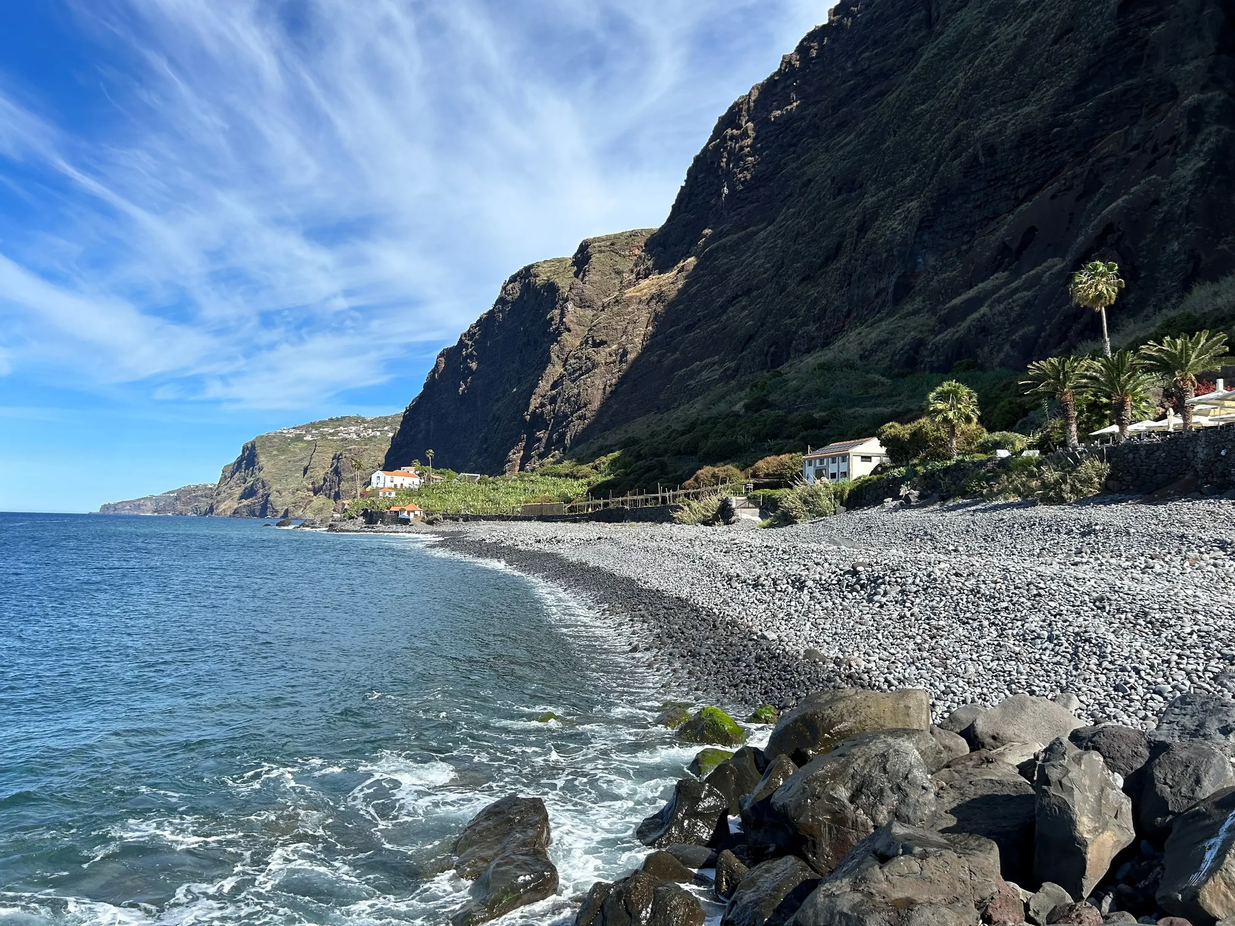 Praia de pedras com ondas do mar, grandes rochas na orla, casas brancas e coqueiros ao pé de um grande rochedo rochoso, sob céu azul com nuvens finas.