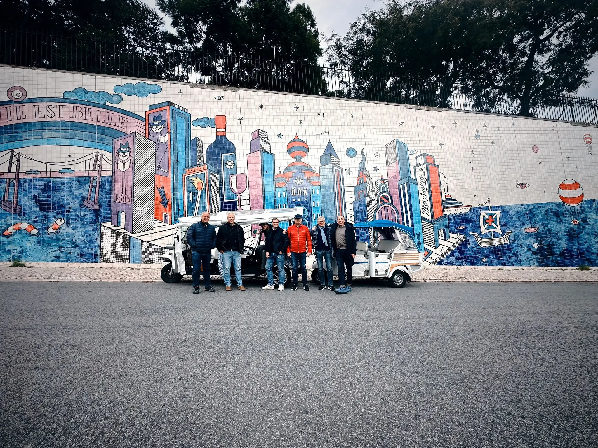 Cinco homens com jaquetas embarcando em tuk-tuks antigos na frente de um mural colorido que retrata um cenário urbano futurista com prédios altos, barcos e elementos de ficção científica.