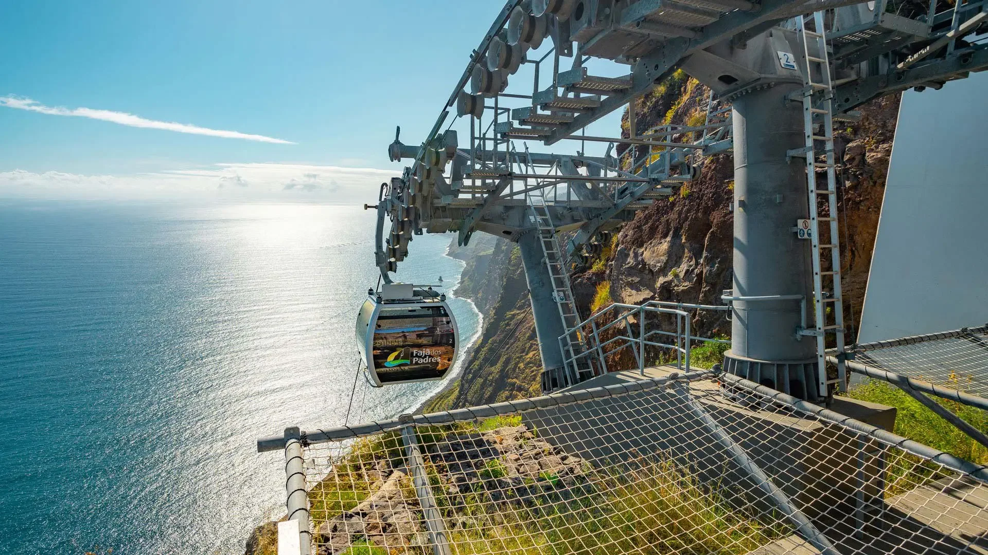 Teleférico em um parque costeiro, com vista para o oceano e penhascos, incluindo uma cabine do teleférico com a inscrição "Fajados Padre".