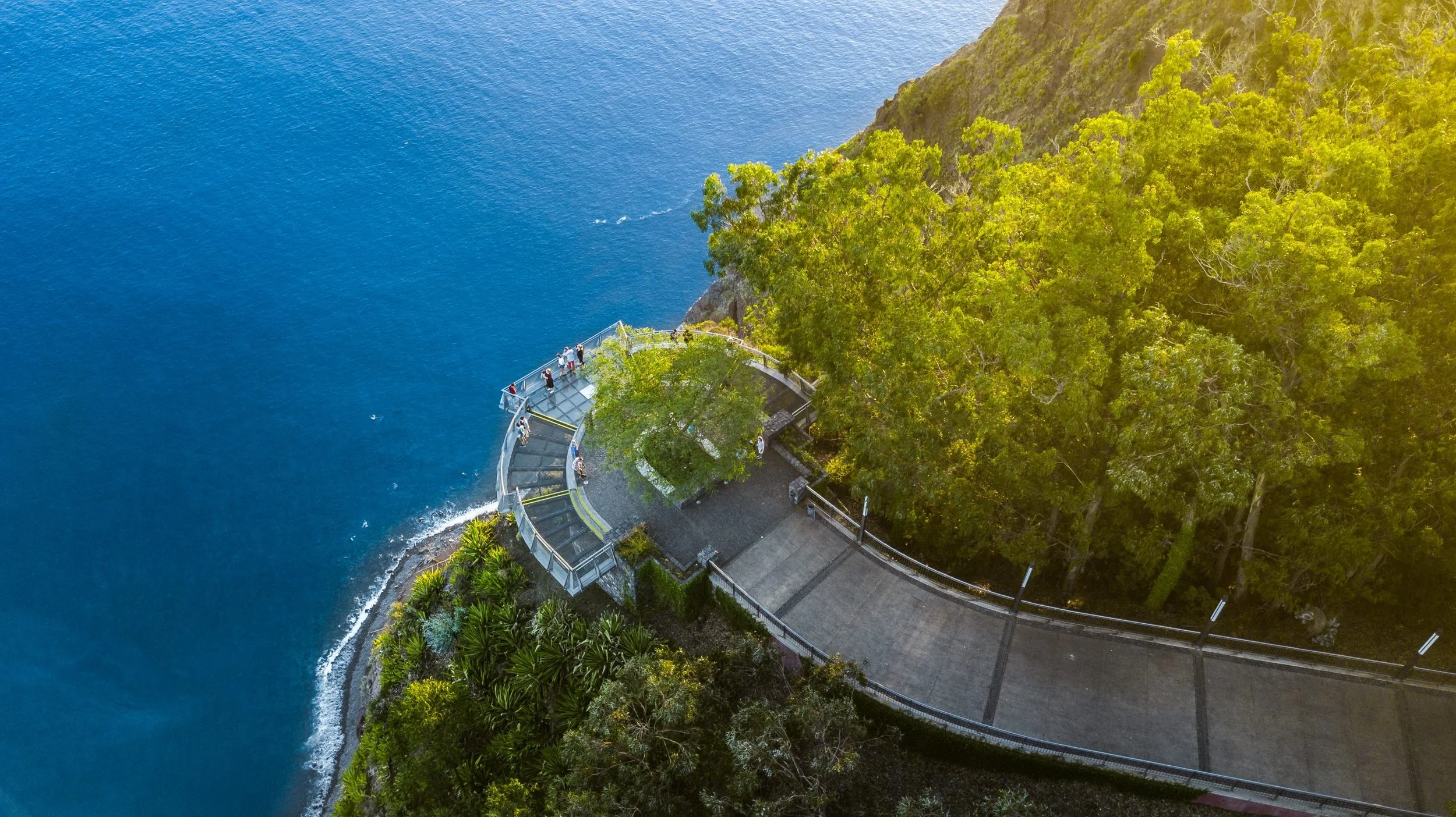 Passeio com vista para o oceano, com pessoas na varanda de um ponto de observação em uma área com árvores verdes, na encosta de uma montanha.