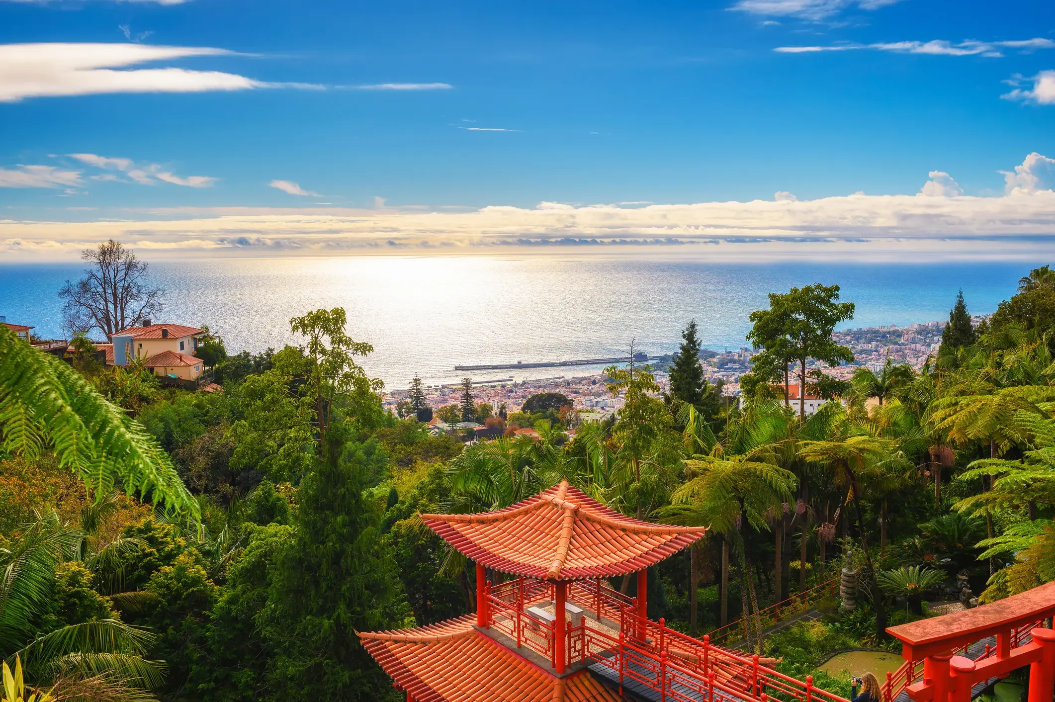 Vista de um jardim com árvores e um pavilhão vermelho em estilo oriental, com o mar e uma cidade ao fundo sob um céu azul com nuvens.