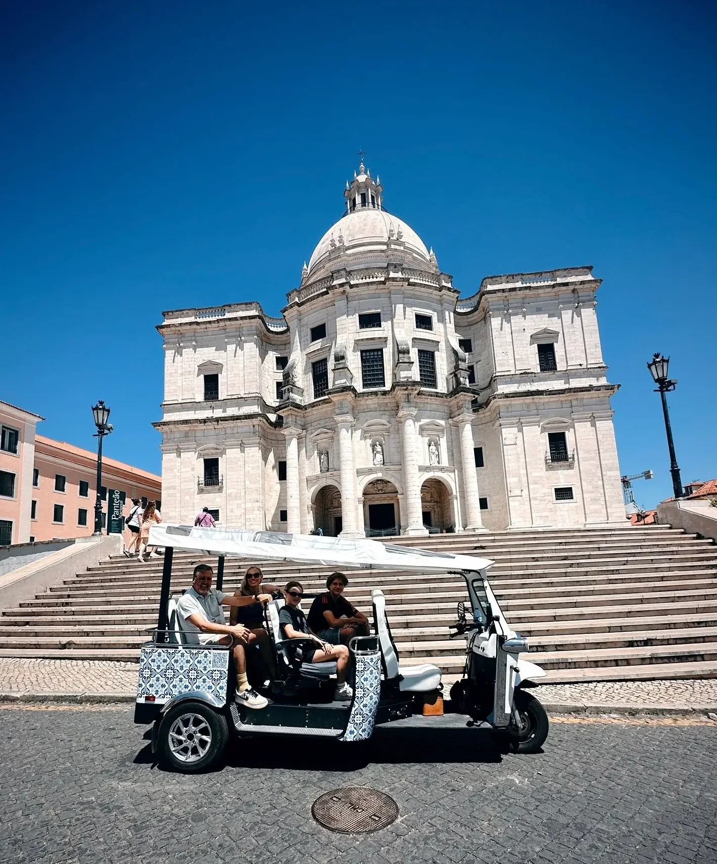 Um grupo de quatro pessoas andando de TUKTUK elétrico na frente de um edifício histórico, que parece uma igreja, com degraus altos e um grande domo, sob céu azul.