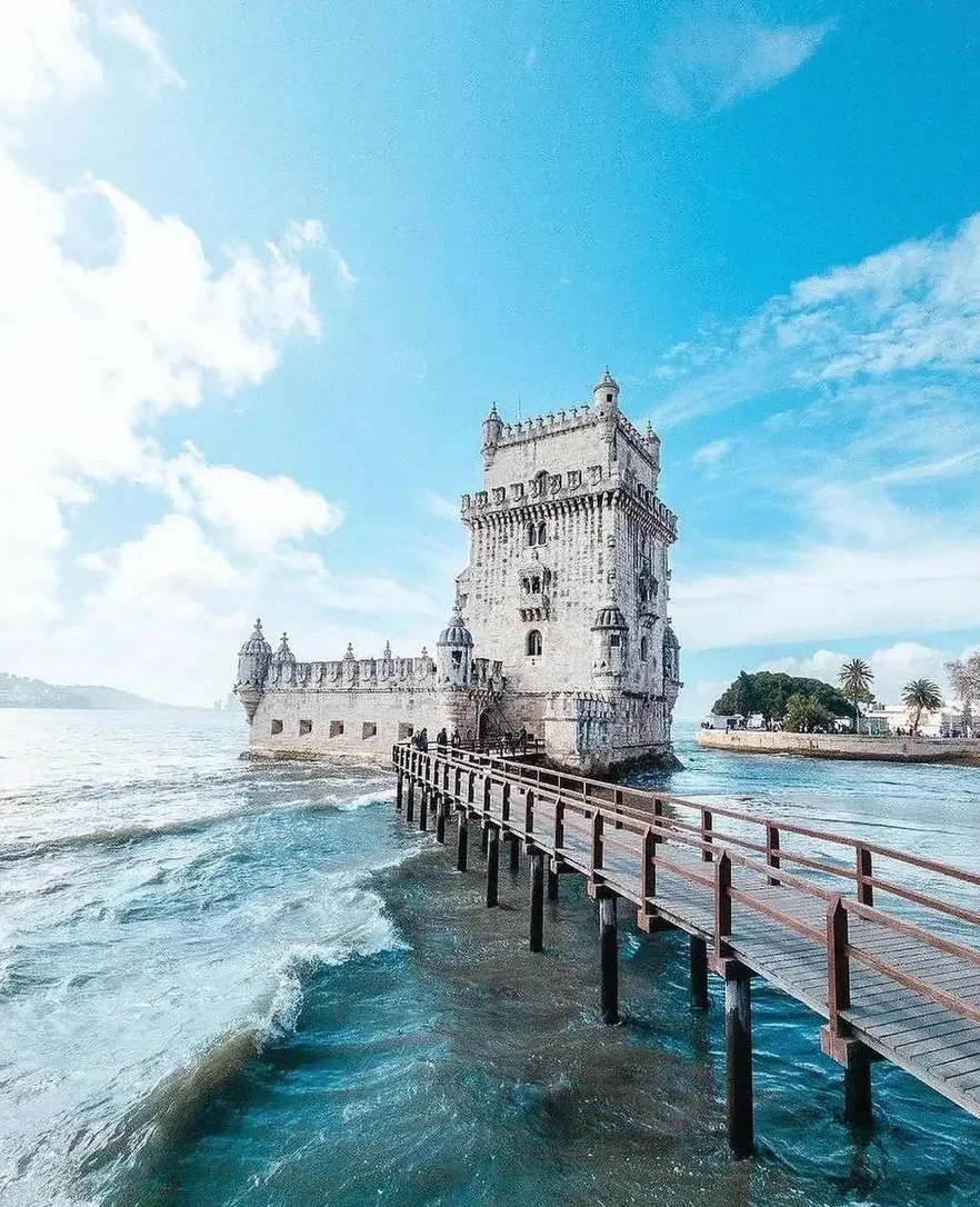Castelo sobre o mar com ponte de madeira levando até ele, céu azul com nuvens
