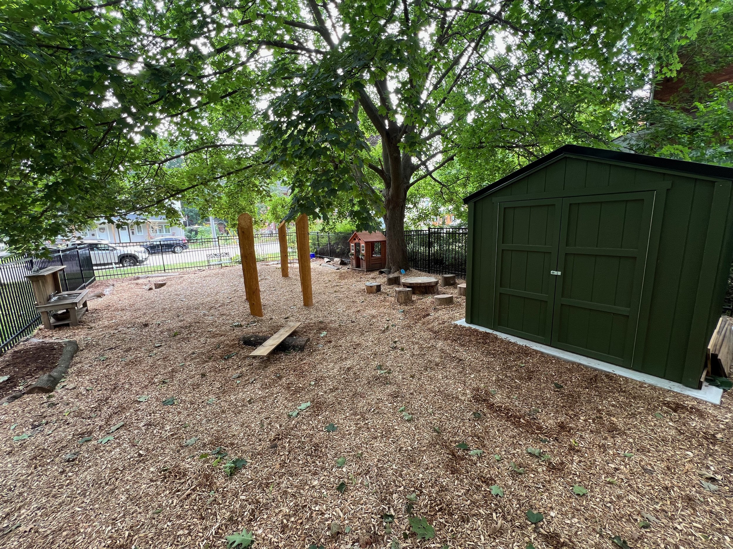 Natural playground with a large leafy tree, a green shed, a small playhouse, and a fenced area with parked cars in the background.
