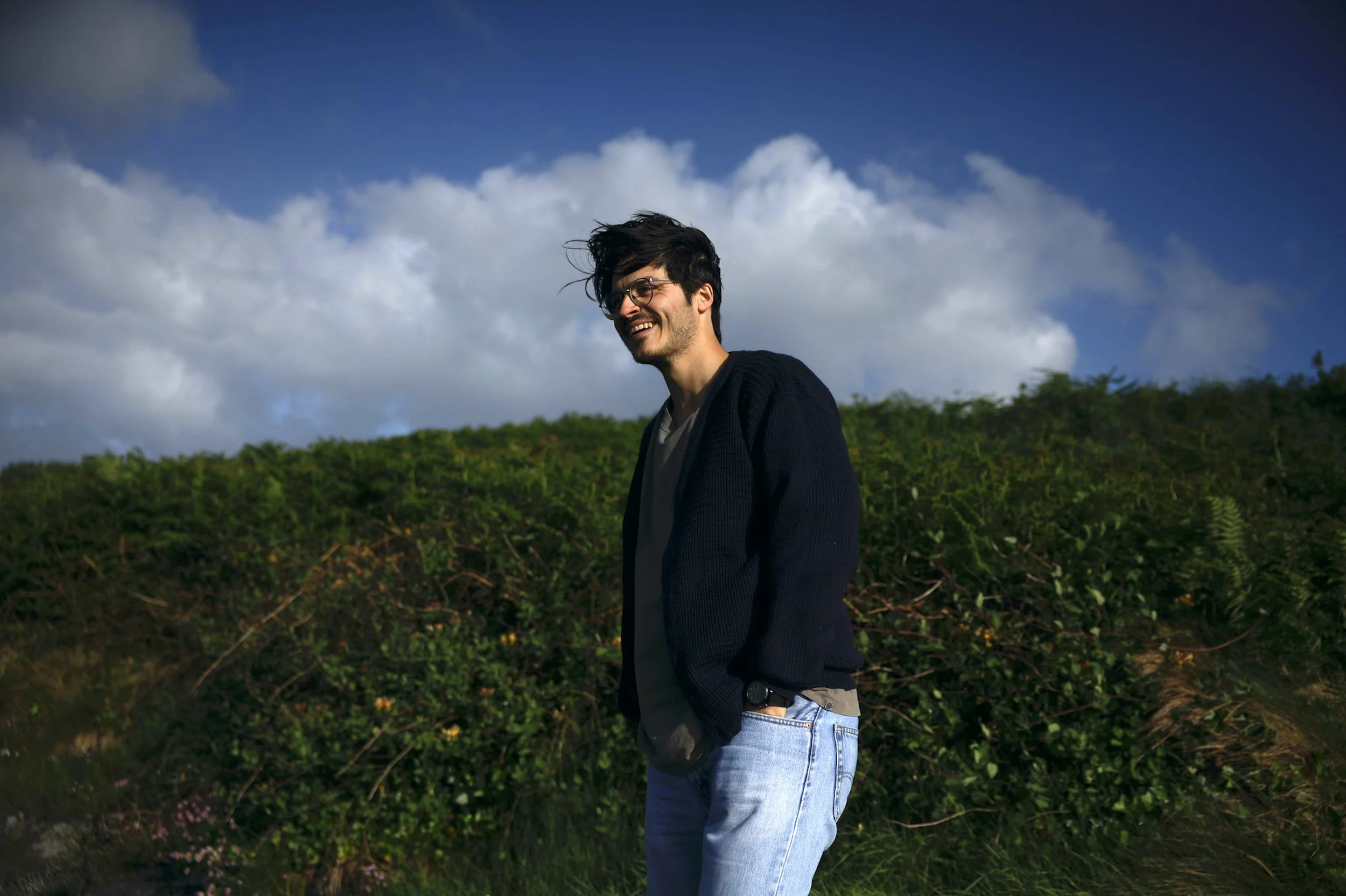 A young man with dark hair, glasses, and casual clothing, smiling and standing outdoors in front of green bushes with a partly cloudy sky overhead.