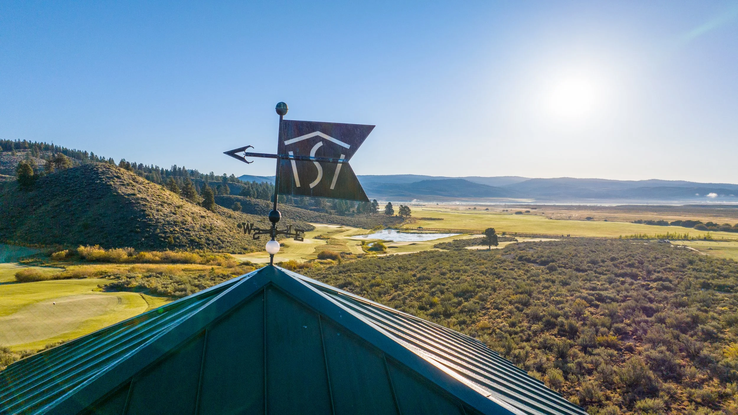 A weather vane with a black arrow and directional markers, mounted on a teal-colored roof, overlooking a landscape of hills, fields, and a river under a bright blue sky.