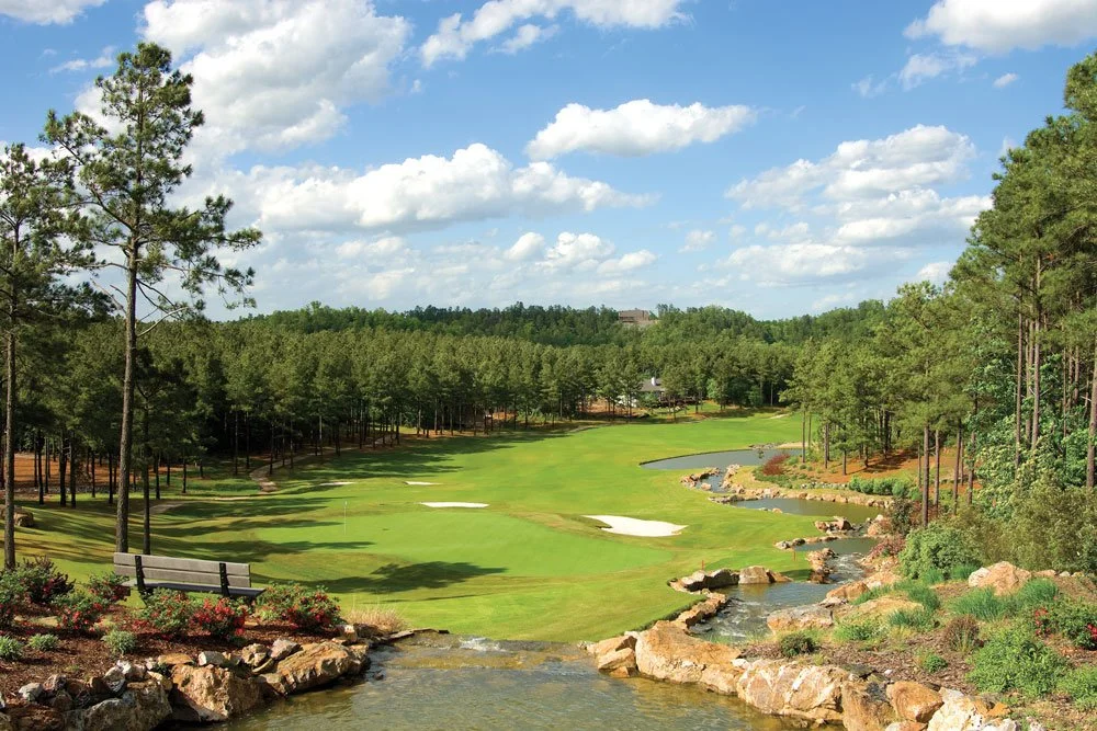 A scenic golf course with green fairways, sand bunkers, and a small stream flowing through the landscape, surrounded by tall trees under a partly cloudy sky.