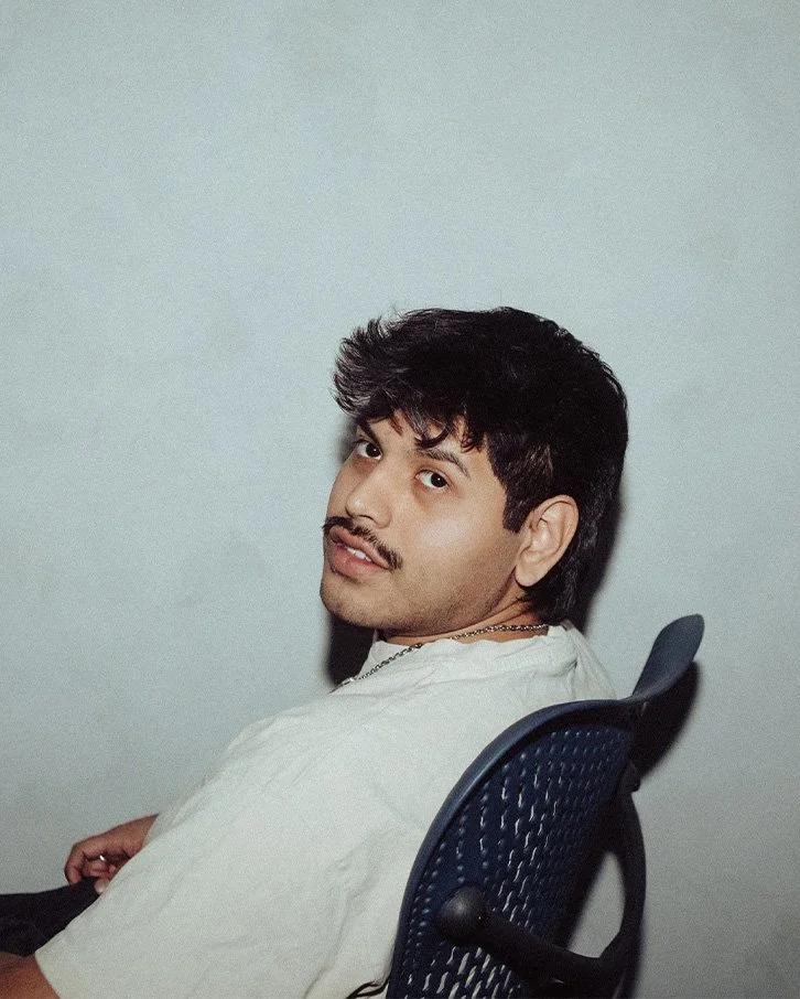 A young man with dark, wavy hair and a mustache, sitting on a blue office chair, looking over his shoulder at the camera against a plain light-colored background.