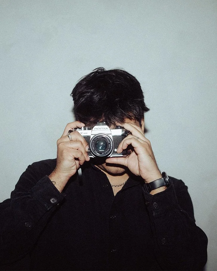 Person with dark hair taking a photo with a vintage film camera against a plain light-colored wall.