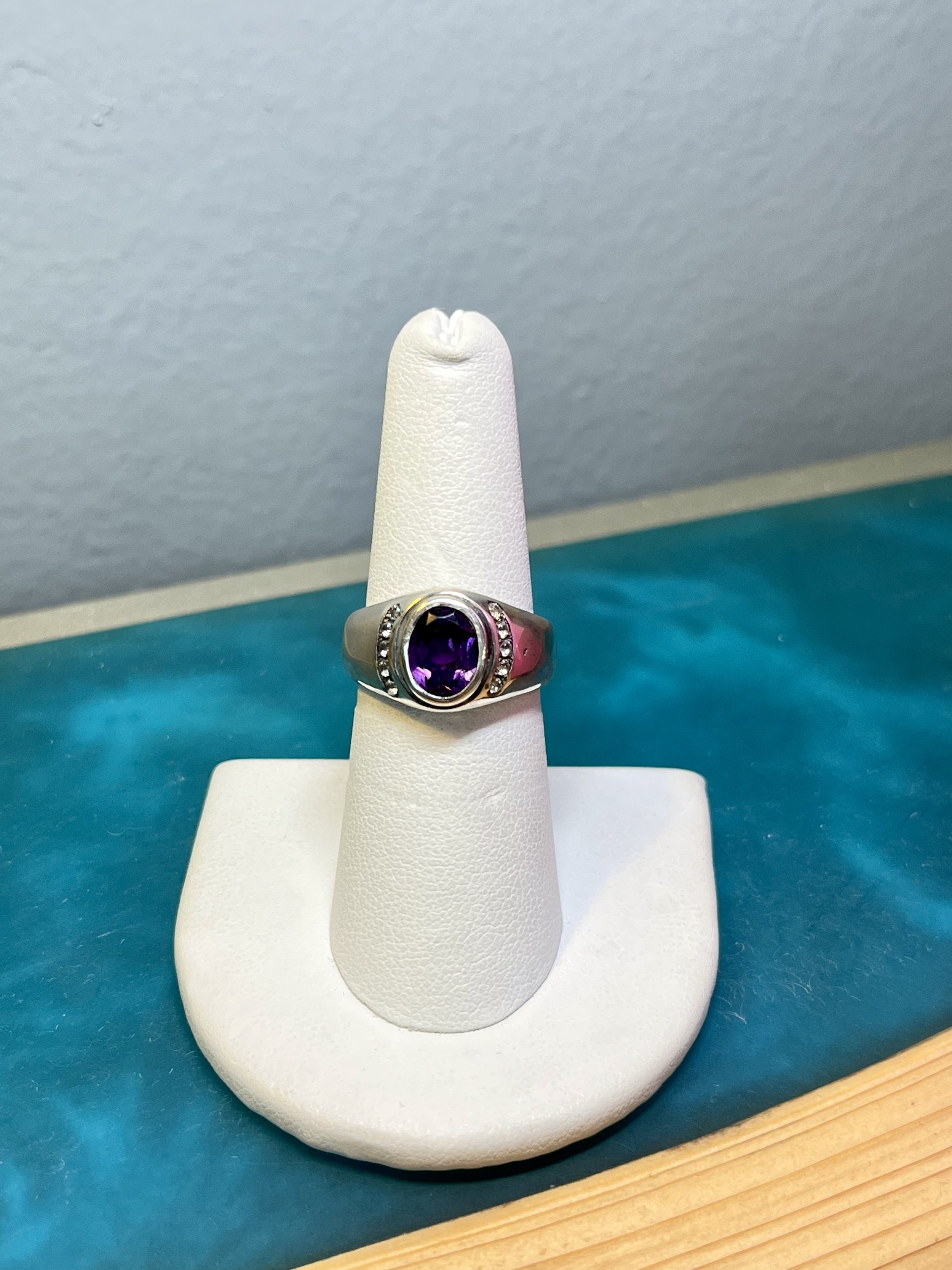 Silver ring with a large oval purple gemstone and small surrounding diamonds, displayed on a white ring holder.