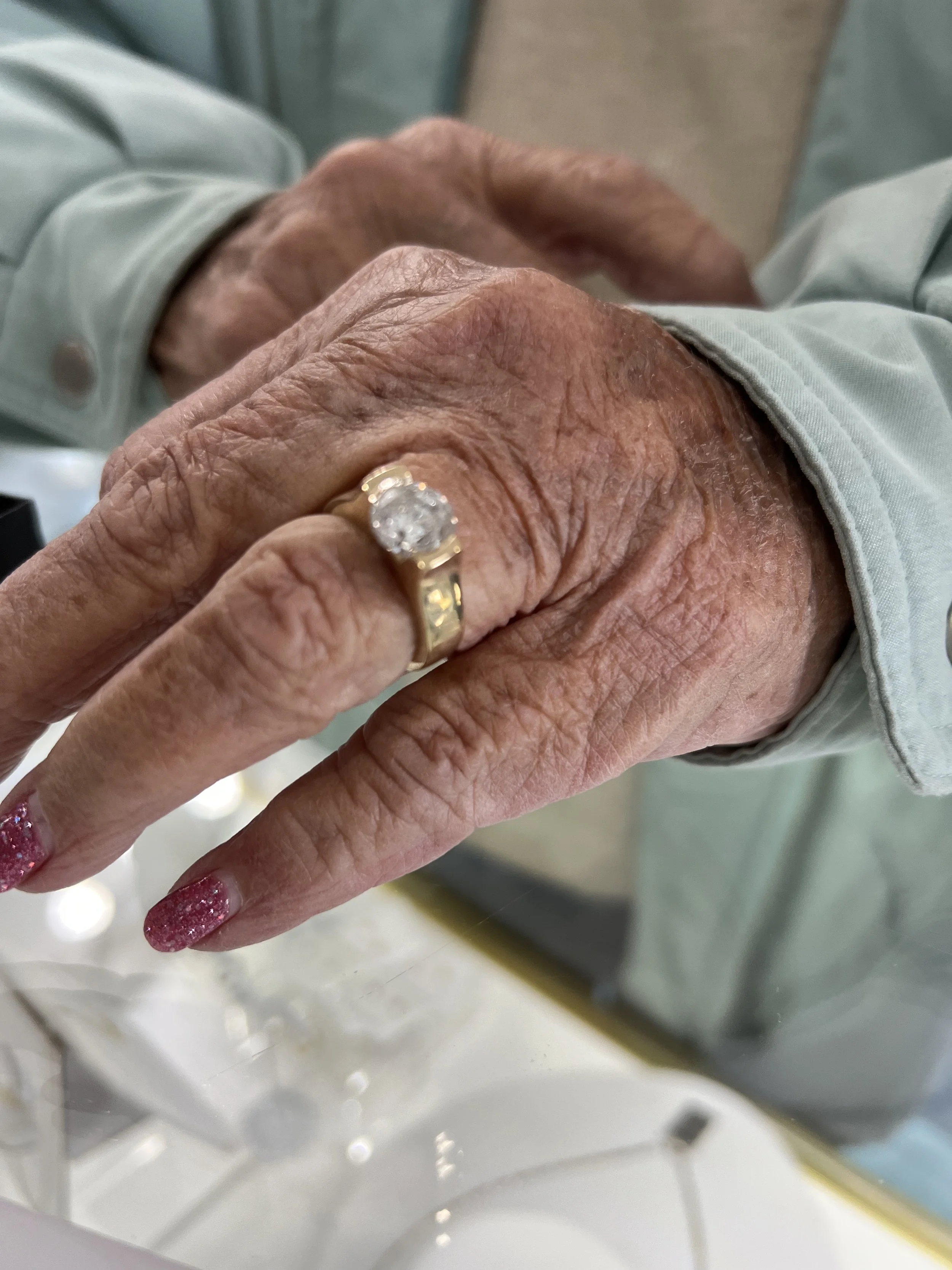 Close-up of elderly person's hand with a gold ring featuring a large clear gemstone, resting on another person's hand, with pink glittery nail polish and wrinkled skin.
