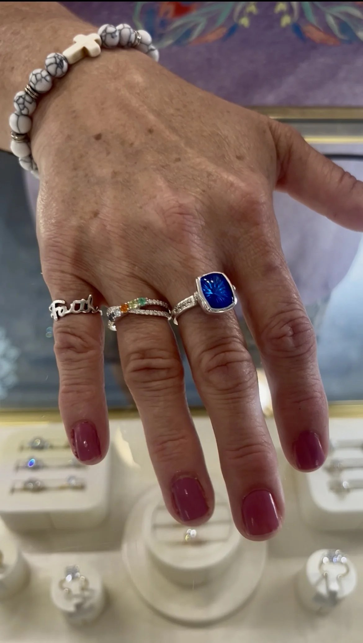 Close-up of a woman’s hand with multiple rings, including a large blue gemstone ring, a ring with the word 'Faith', and a ring with colorful stones. She is wearing beaded bracelets on her wrist. In the background, jewelry displays with various rings 