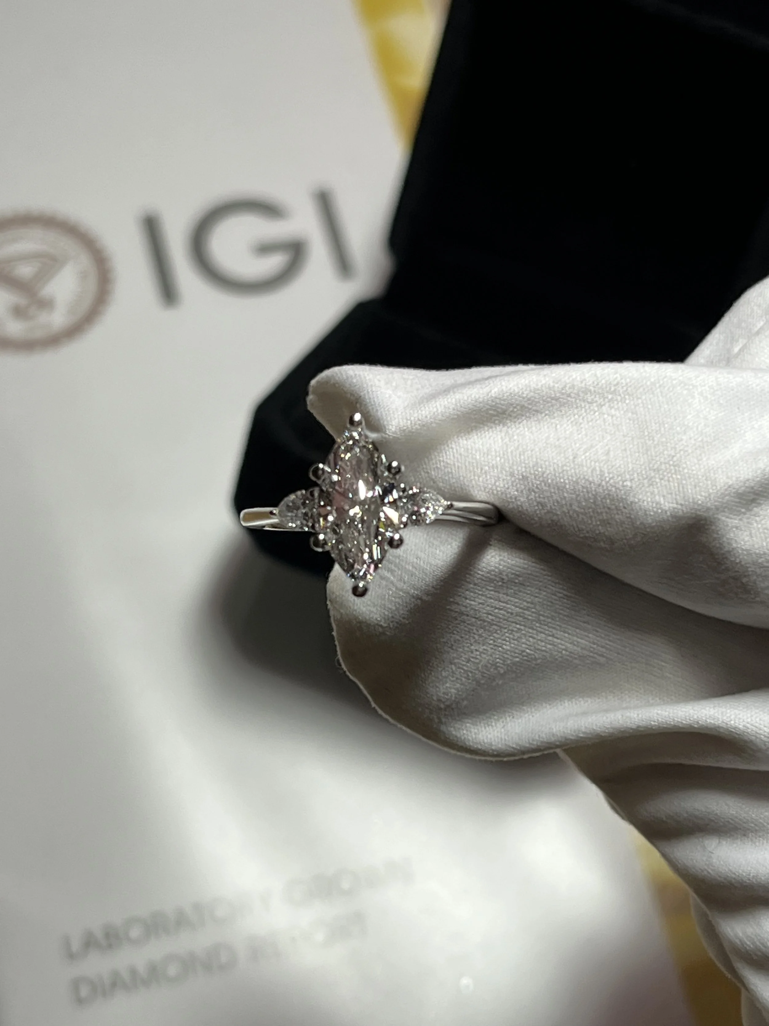 A diamond ring with a marquise-shaped center stone surrounded by smaller diamonds, held in a white glove against a white background with text and logo.