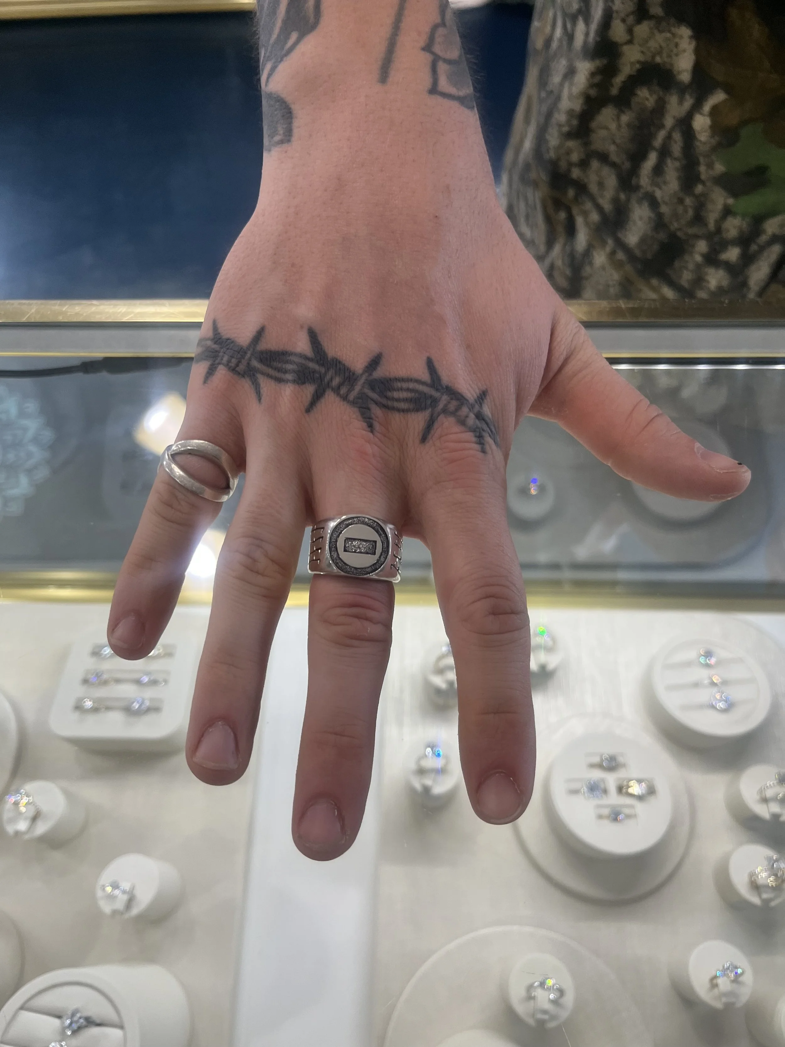 A person's hand with a barbed wire tattoo on the wrist, multiple rings on fingers, and a display case with jewelry in the background.