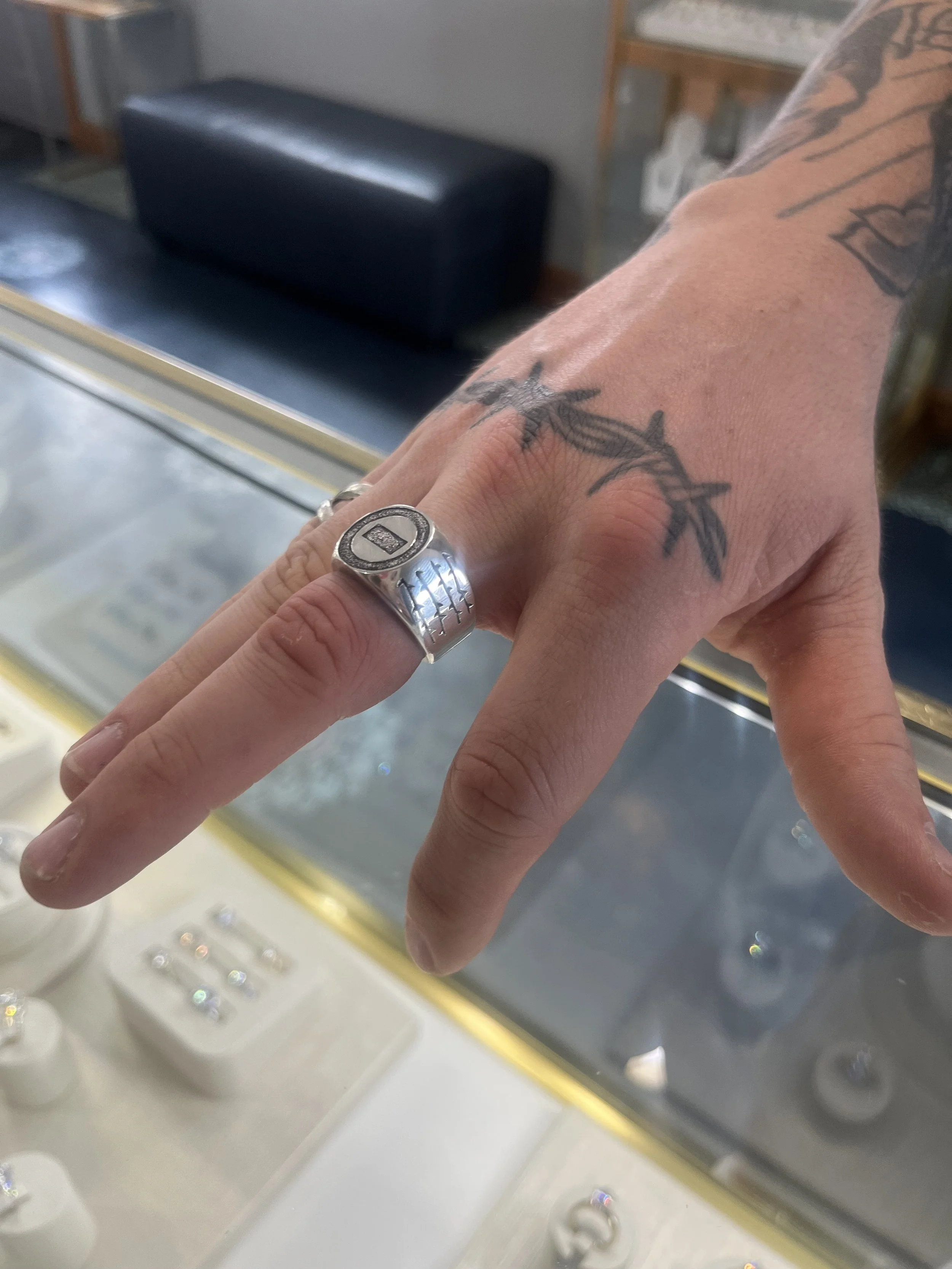 A hand with a silver ring on the middle finger and tattoos on the back and side of the hand, resting on a glass display case with jewelry inside.