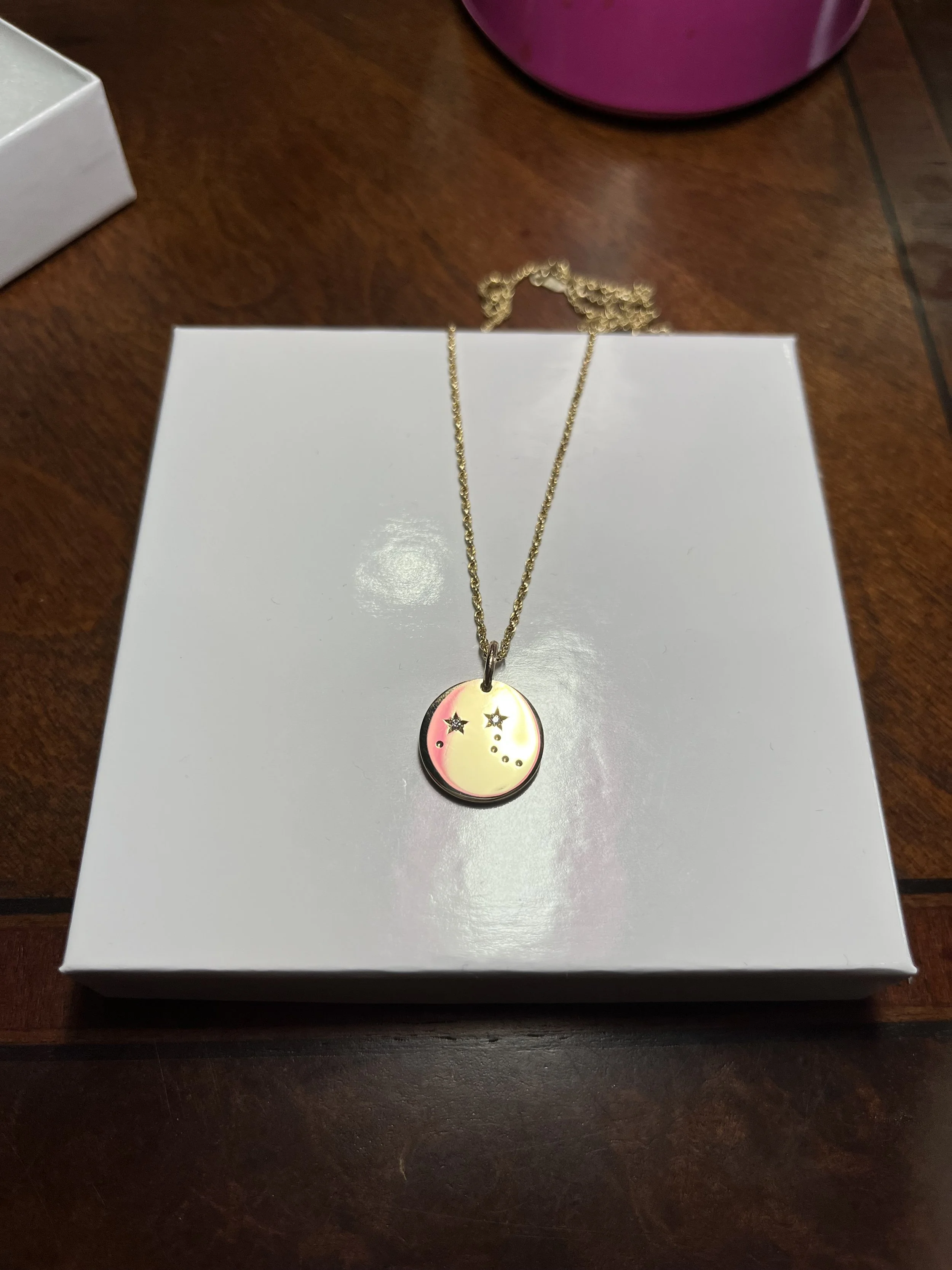 Gold necklace with a round pendant featuring a star and dot design, placed on a white box.