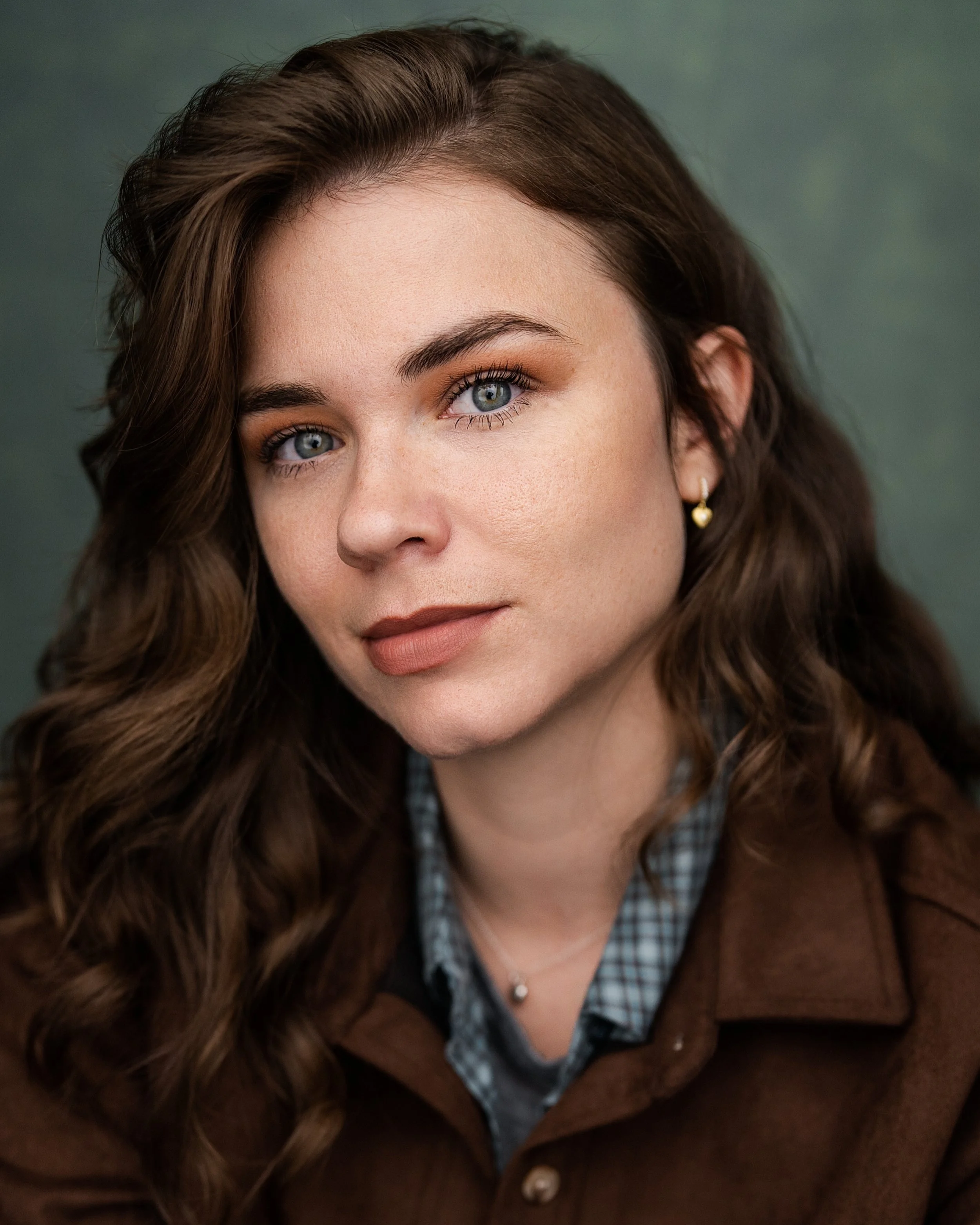 Close-up portrait of a woman with wavy brown hair, blue eyes, and light makeup, wearing a brown jacket, checked shirt, and small gold earrings, against a green background.