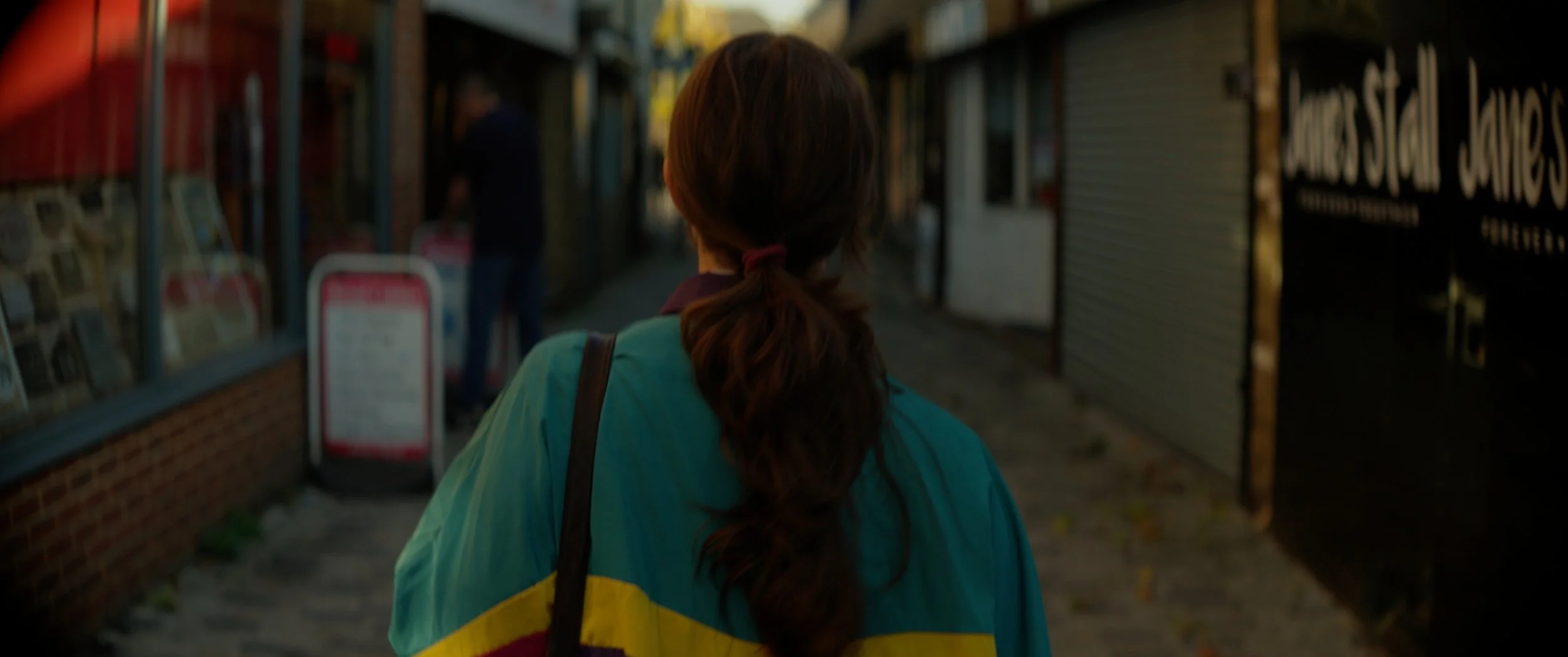 A woman with long brown hair tied in a ponytail, seen from behind, walking down a city sidewalk during daytime, wearing a colorful jacket with blue, yellow, and red sections. There are storefronts and people in the background.