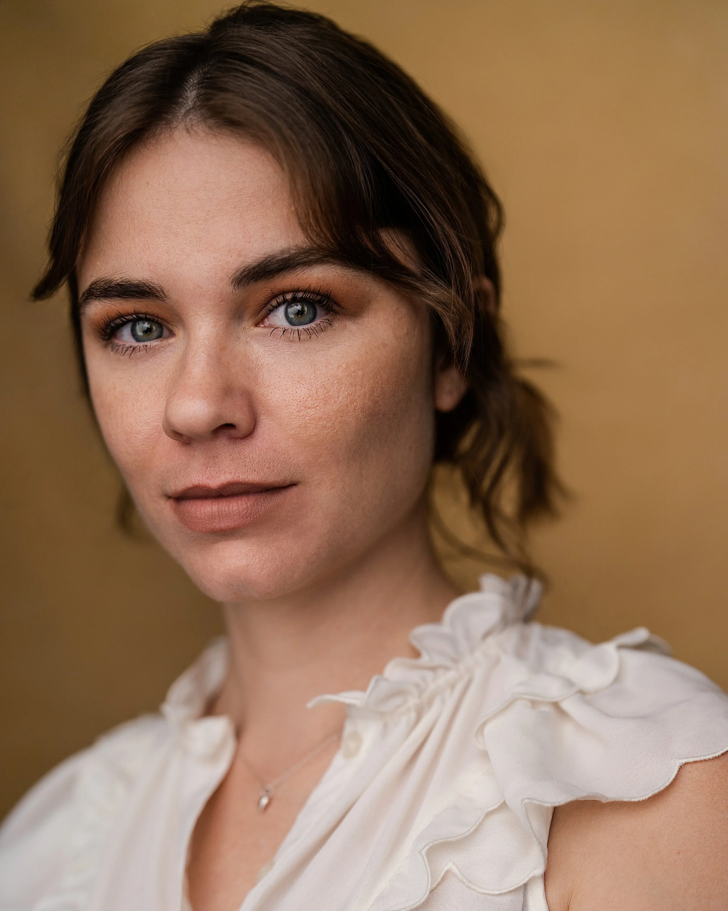 Close-up portrait of a young woman with short brown hair and blue eyes, wearing a white ruffled blouse and a delicate necklace, looking at the camera with a neutral expression.