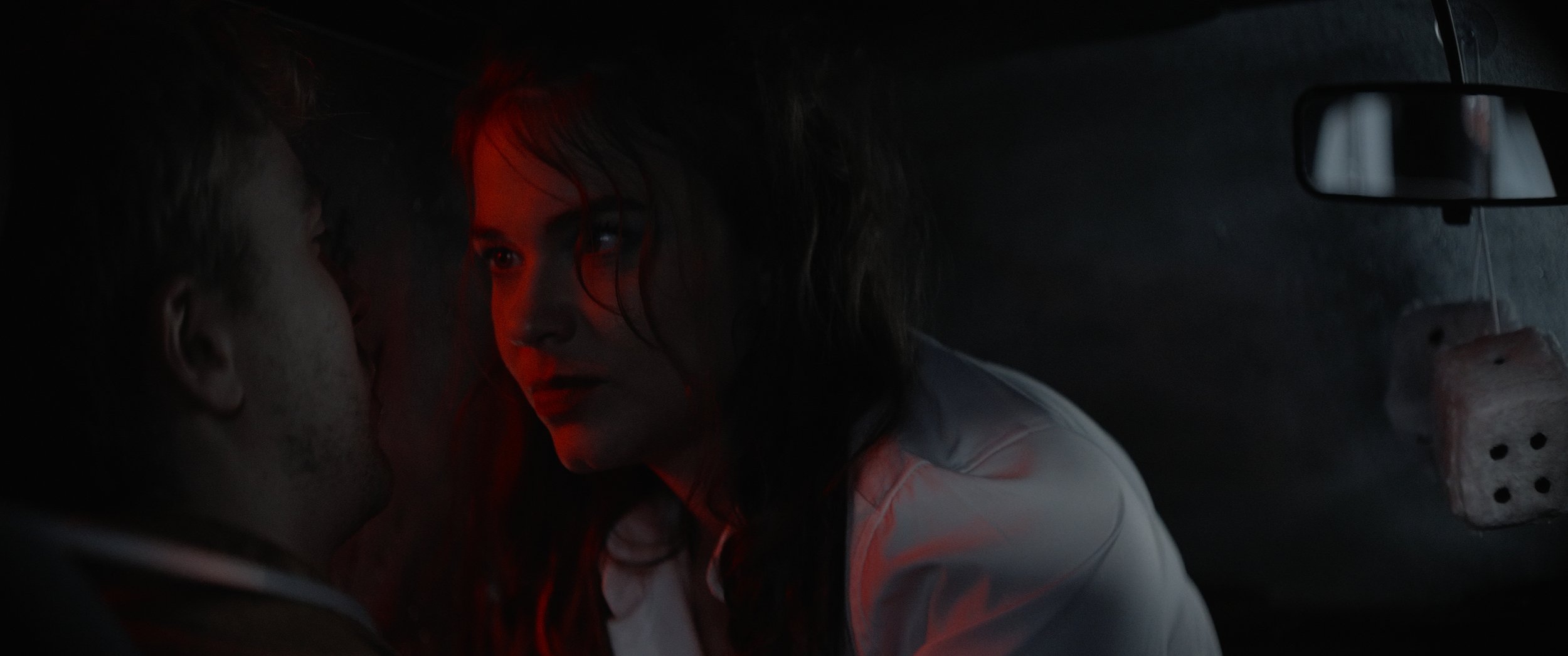 A woman and a man face each other inside a dark car, illuminated by red lighting, with a large dice hanging from the rearview mirror.