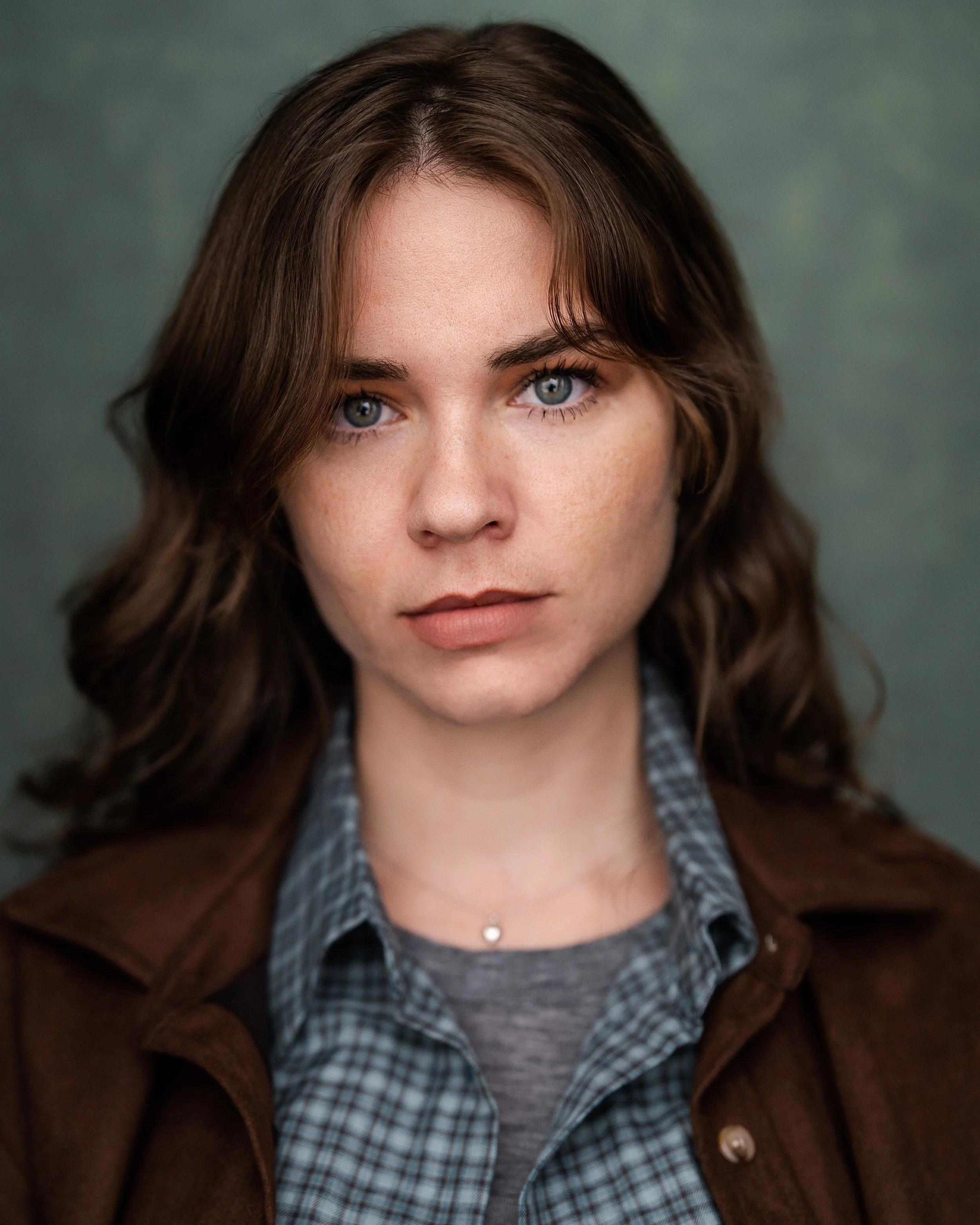 Close-up portrait of a woman with brown, wavy hair and blue eyes, wearing a checkered shirt, a gray top, and a brown jacket, against a blurred green background.