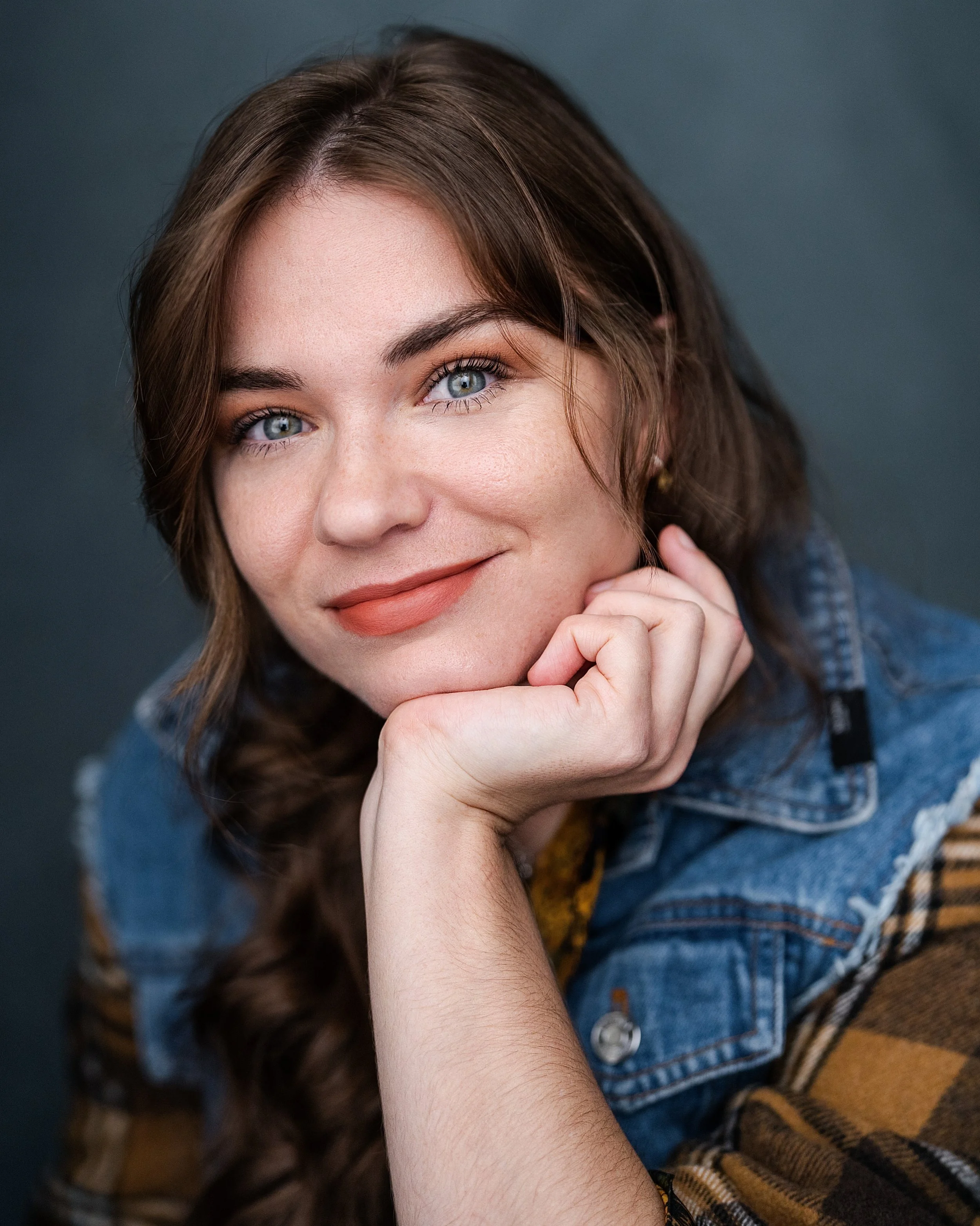 A young woman with long brown hair, blue eyes, and light makeup, smiling with her chin resting on her hand, wearing a denim jacket over a plaid shirt against a dark background.