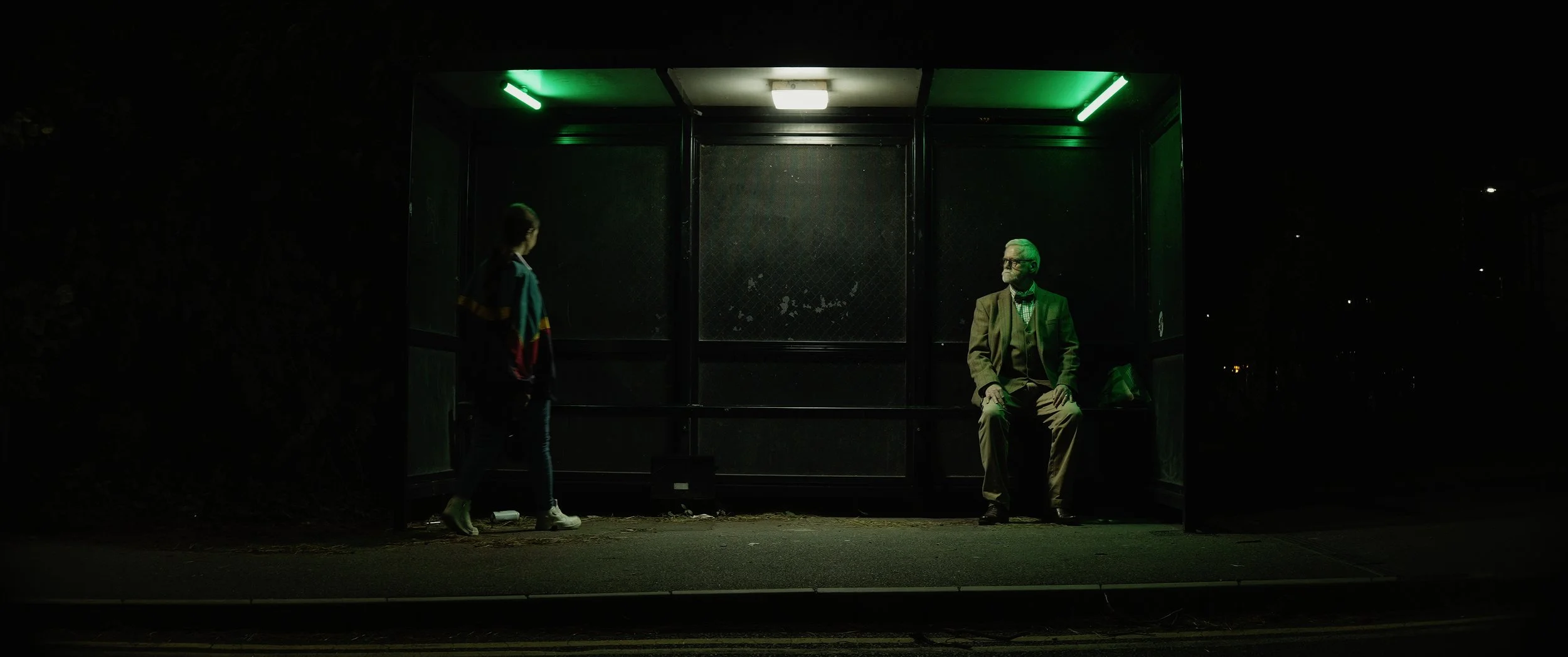Dimly lit bus stop at night with an older man sitting on the bench and a young girl approaching. The bus stop has green lighting and the background is dark with no visible surroundings.
