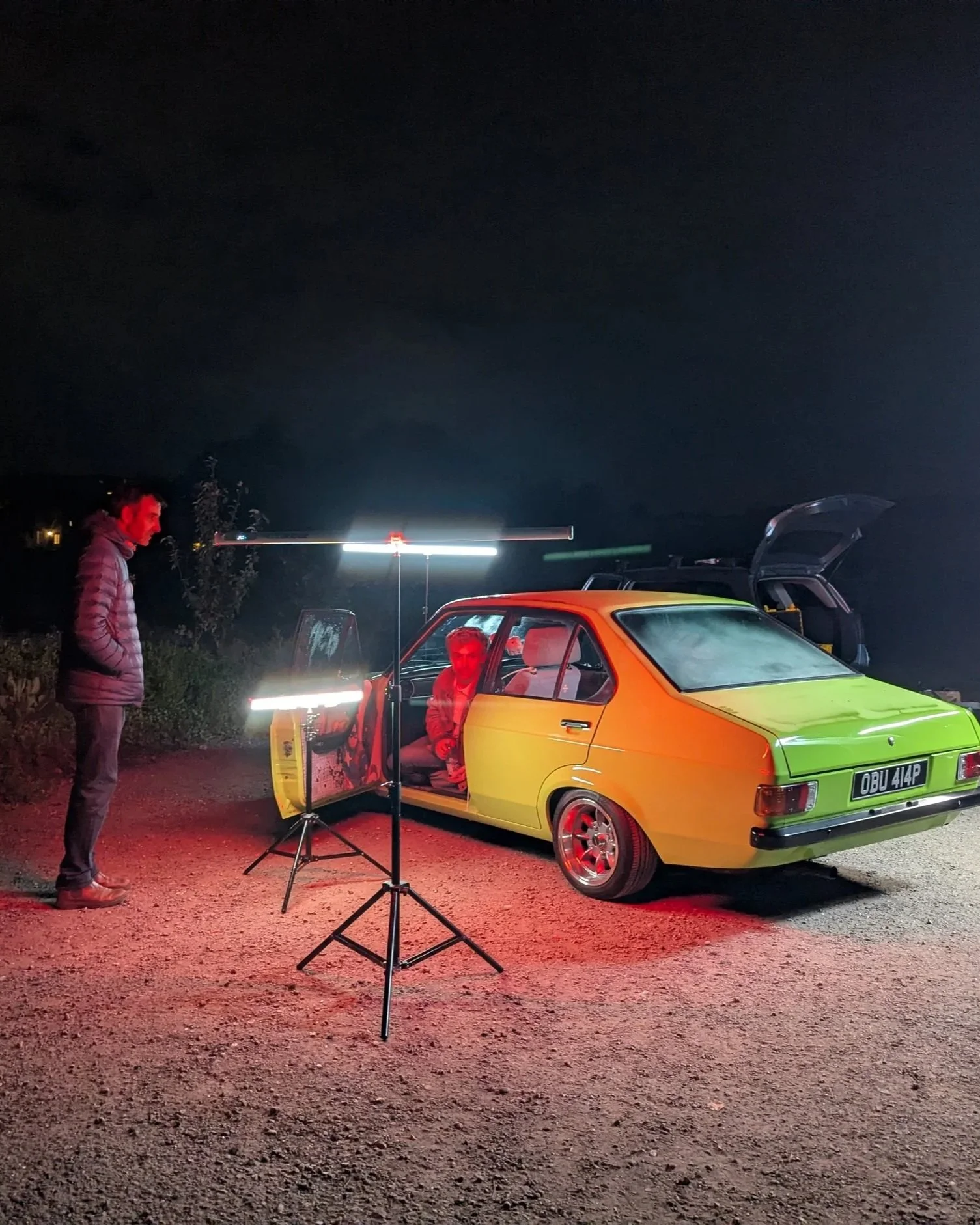 A yellow vintage car with a black license plate parked outdoors at night. Two people are inside the car, and a third person is standing outside near a lighting setup on tripods. The scene appears to be illuminated by red and white lights.