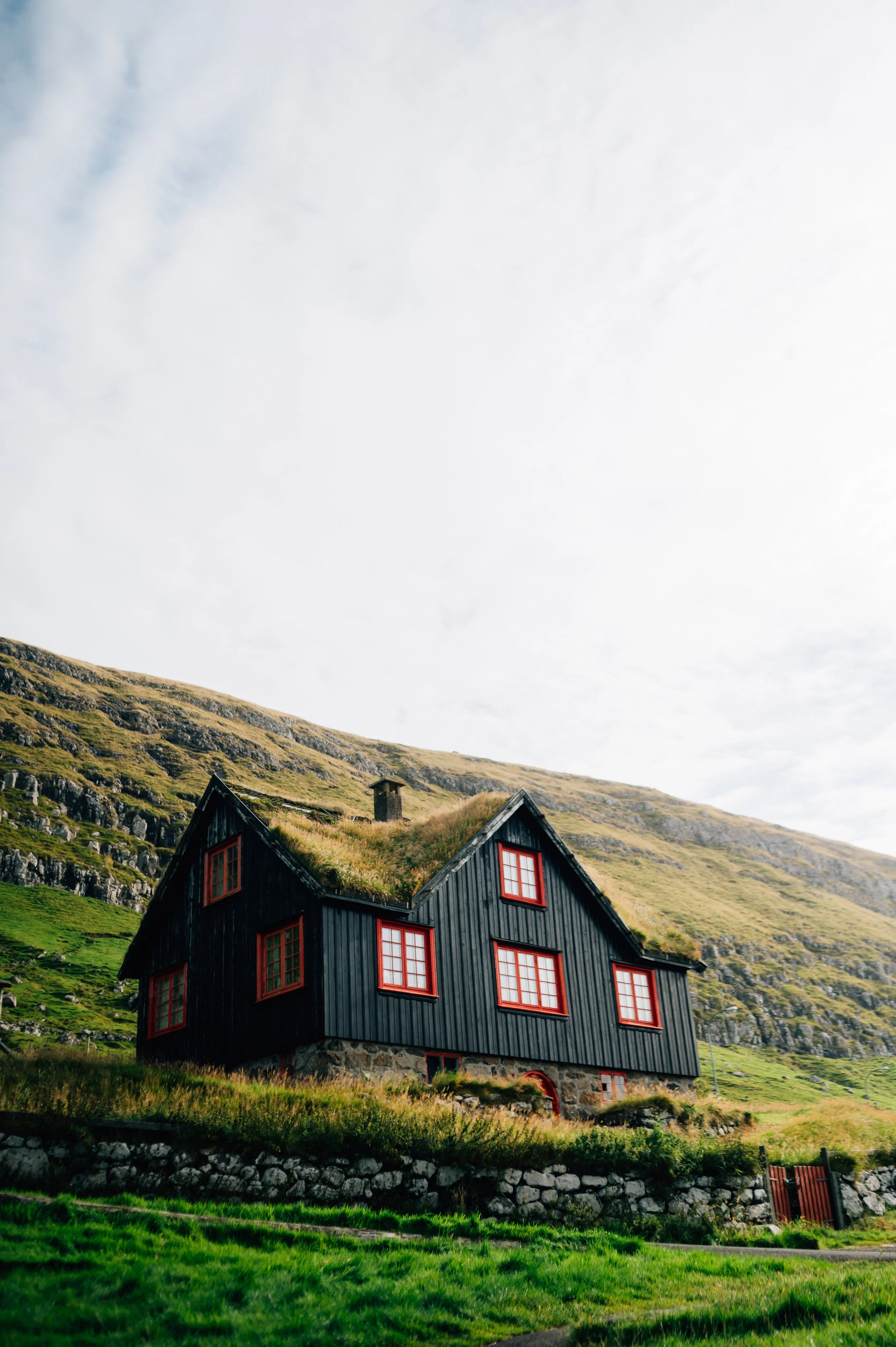 A black house with red windows and a grass roof, sitting on the side of a grassy hill.
