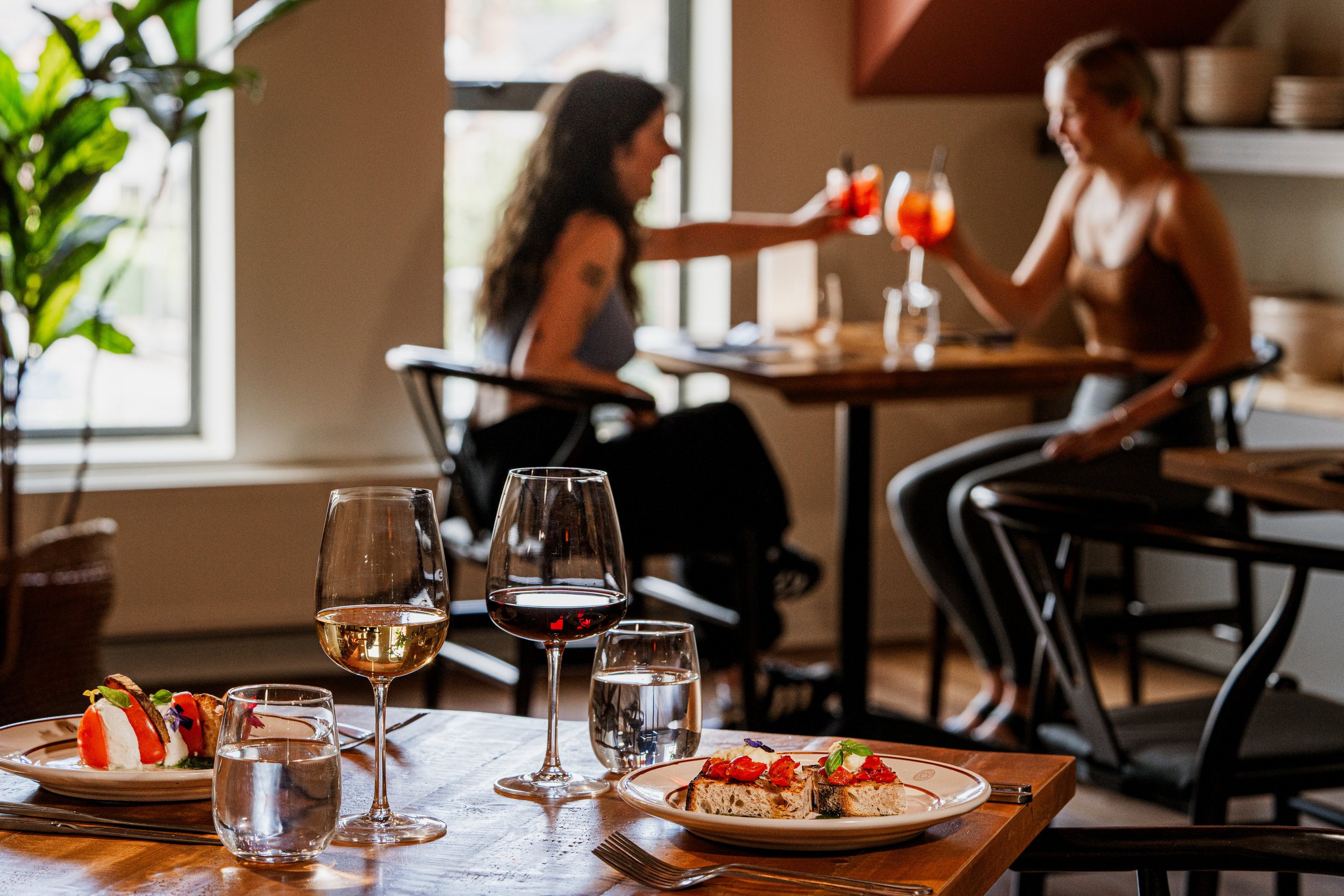 Cozy restaurant with two women toasting drinks in the background; foreground shows plates with appetizers, wine glasses, and water glasses on a wooden table.