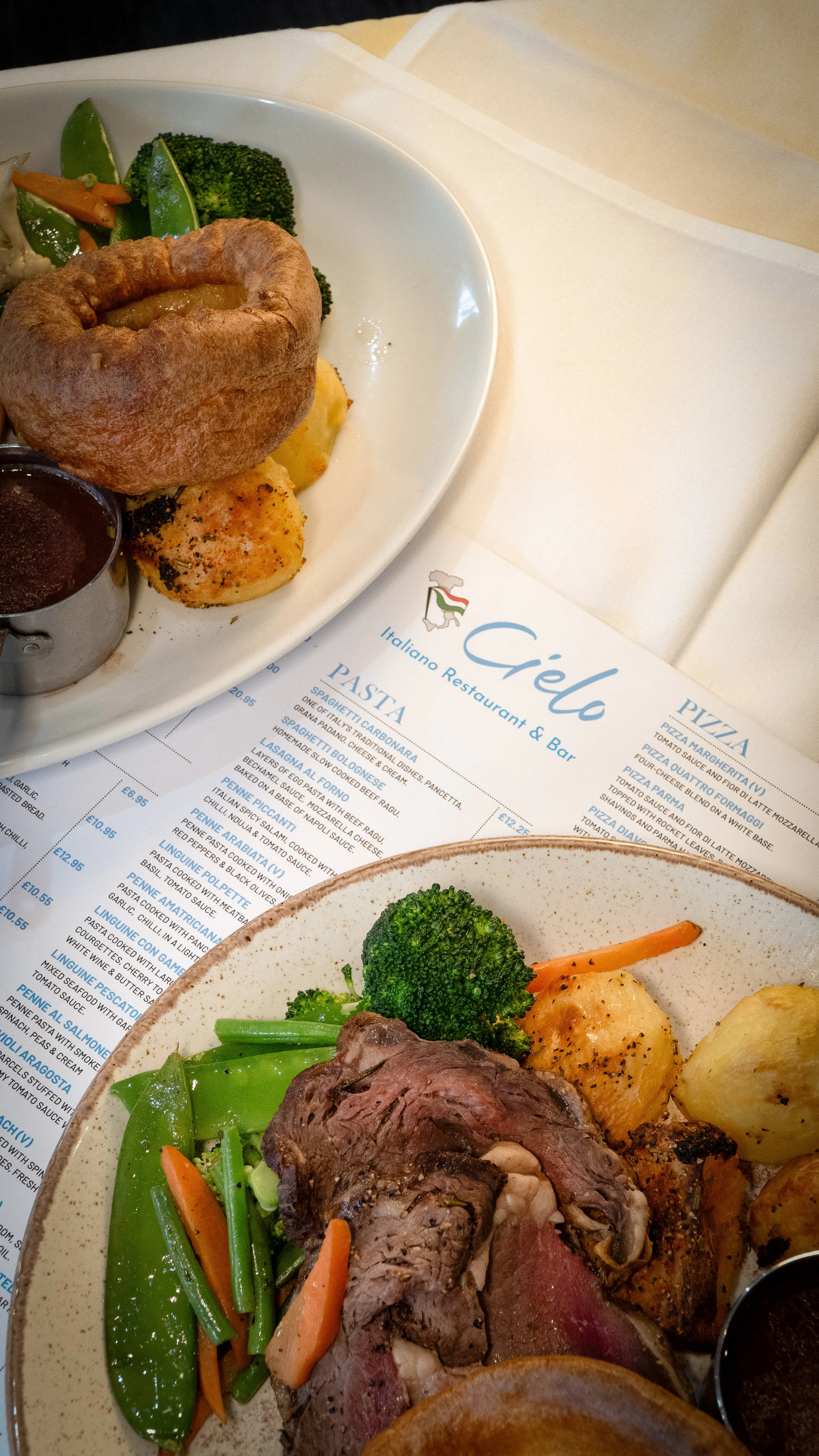 Close-up of a plate with grilled beef, broccoli, carrots, potatoes, and a Yorkshire pudding on a restaurant table with a menu underneath.